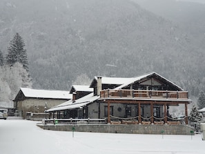 Un amplio refugio con un balcón y un porche cubierto, rodeado de árboles nevados y montañas.