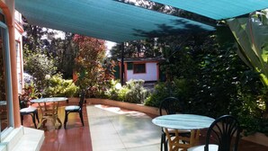 Ein Terrassenbereich im Freien mit Fliesenböden, mehreren Tischen und Stühlen sowie üppiger Bepflanzung.