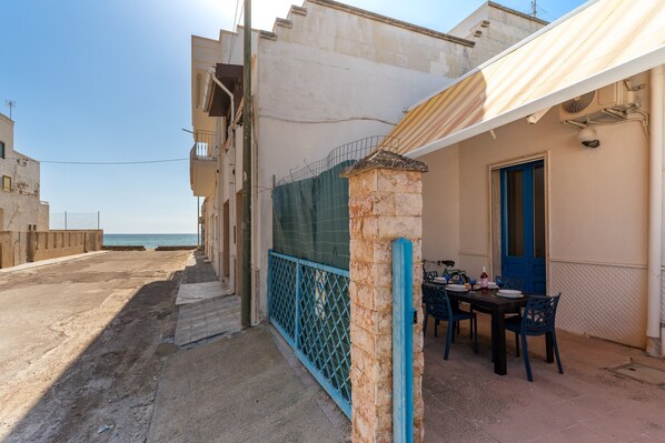 A narrow street with a dining area featuring a table and chairs, a blue door, and a view of the sea.