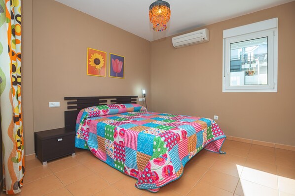 A bedroom with a colorful quilt, a bed, a nightstand, a wall-mounted air conditioner, and a window.