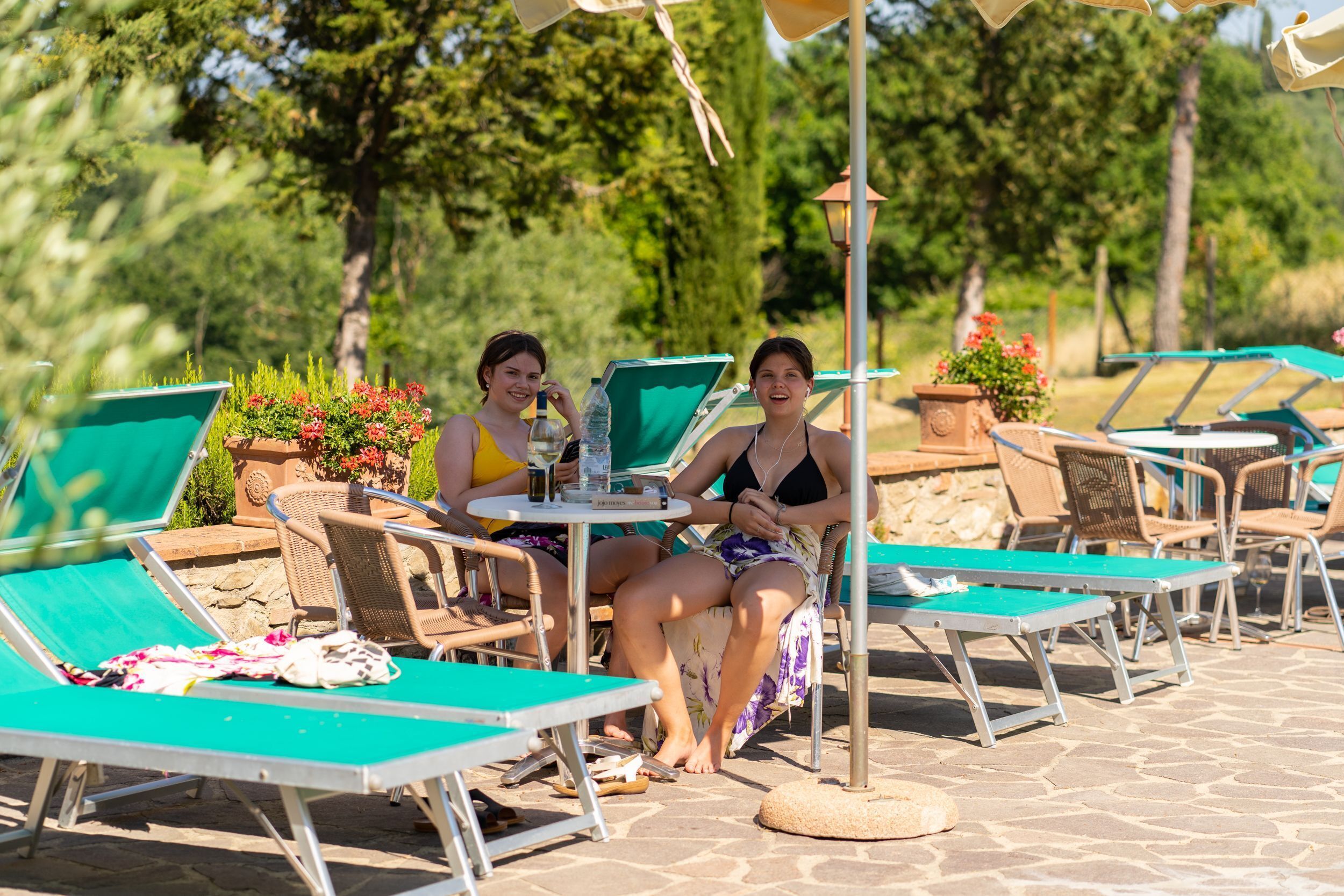 Two people seated at a table with a green umbrella, surrounded by lounge chairs and potted plants.