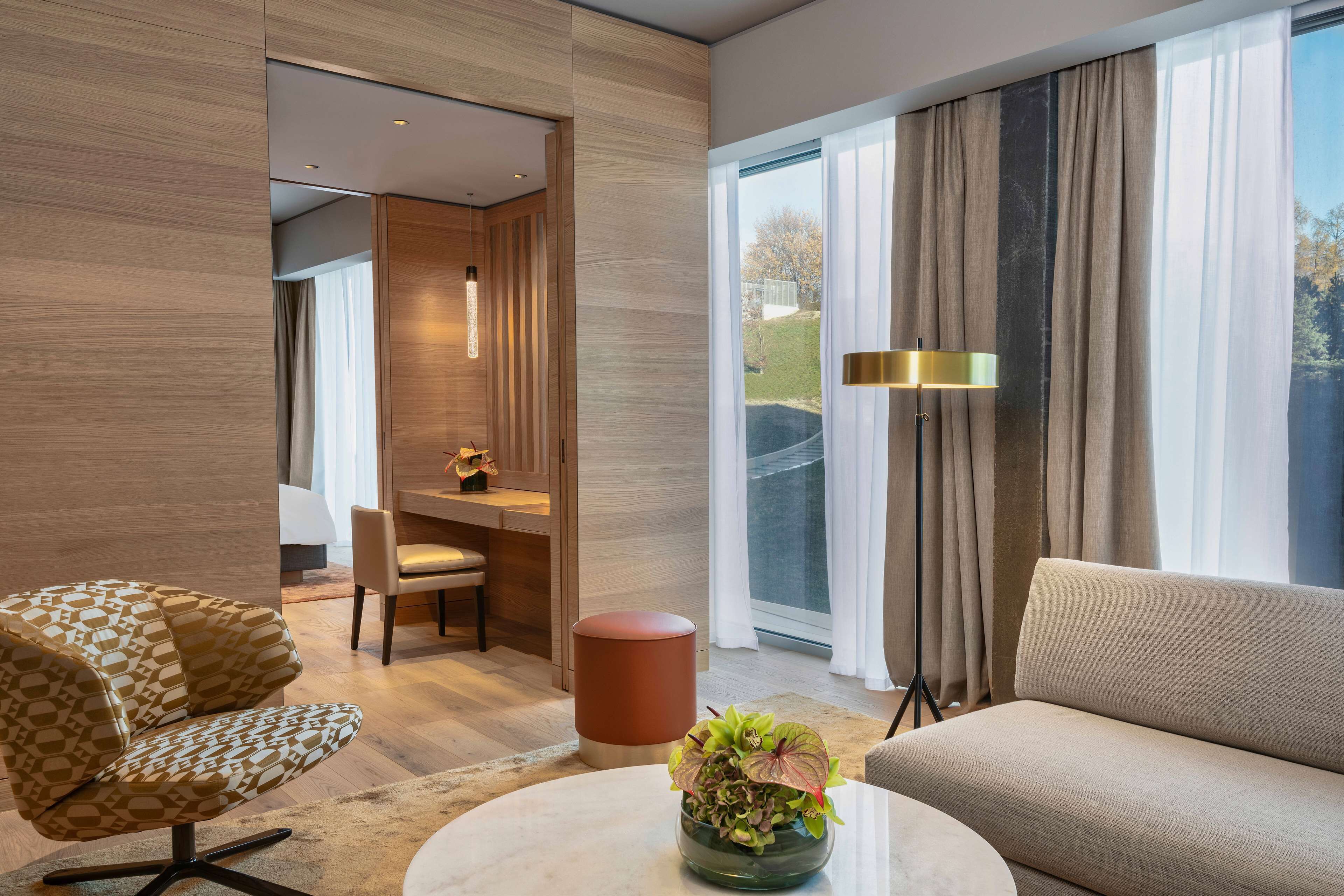 Una habitación de hotel moderna con un ventanal grande, un sofá, una silla, una mesita con una planta y un escritorio con una lámpara.