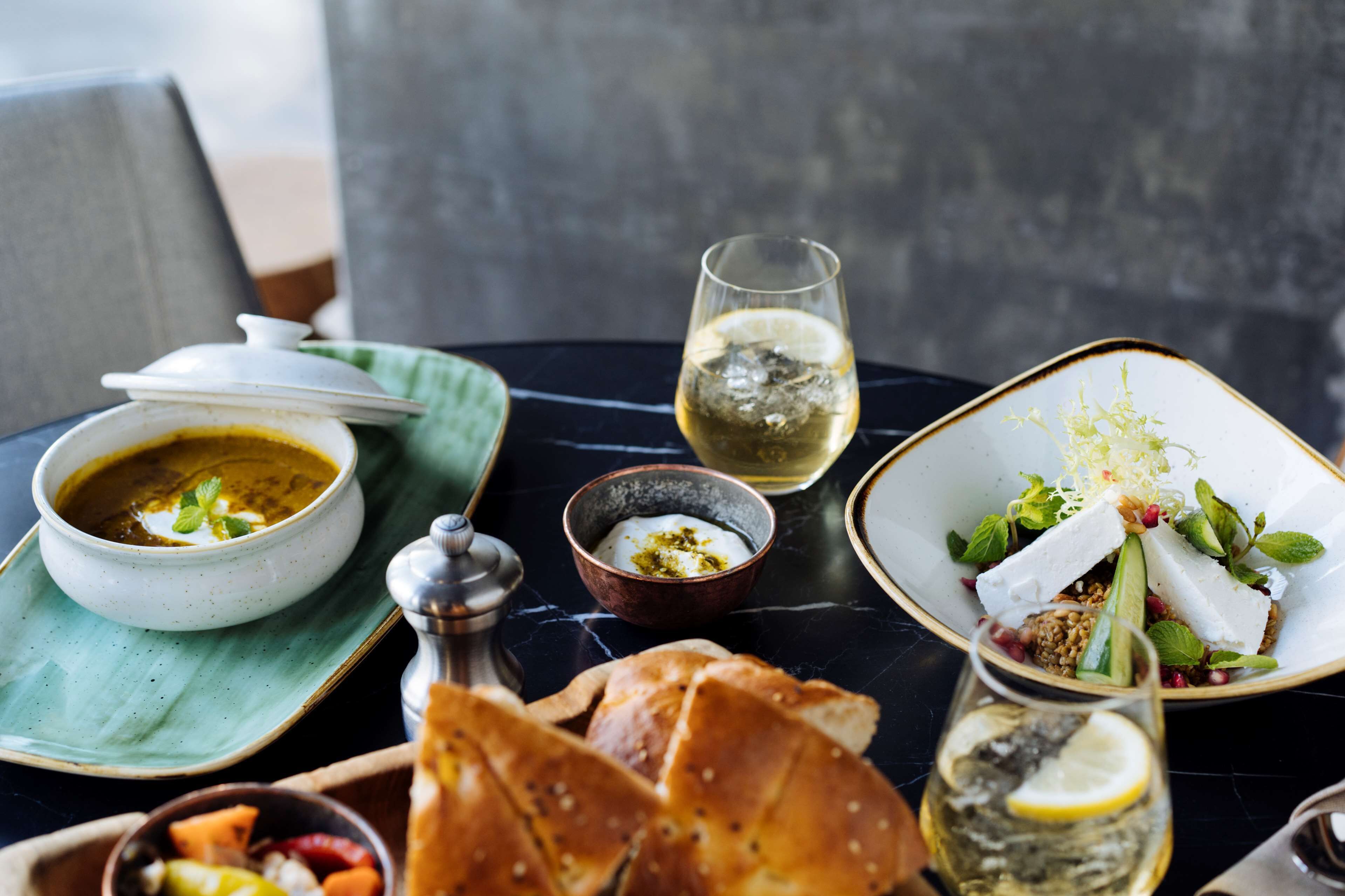 Una mesa con un bol de sopa, una copa de vino blanco, un plato de comida, un pequeño bol de yogur, un vaso de agua y una cesta de pan.