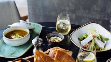 Una mesa con un bol de sopa, una copa de vino blanco, un plato de comida, un pequeño bol de yogur, un vaso de agua y una cesta de pan.
