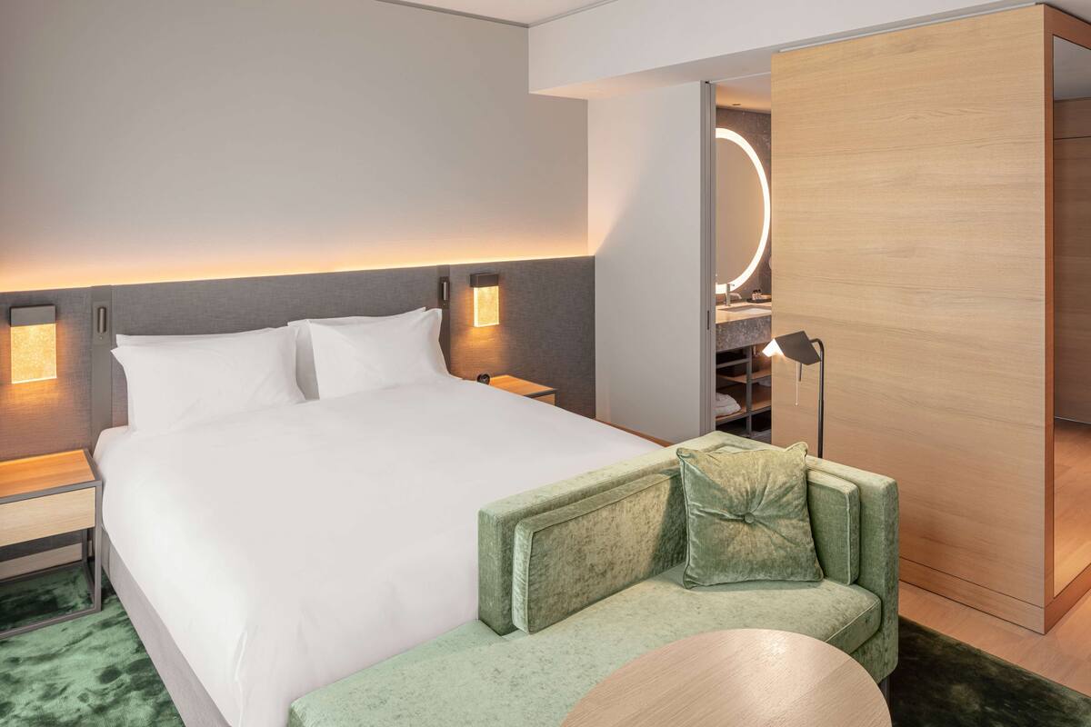 Een moderne hotelkamer met een groot bed, een groene fauteuil, een ronde spiegel en een houten garderobe.