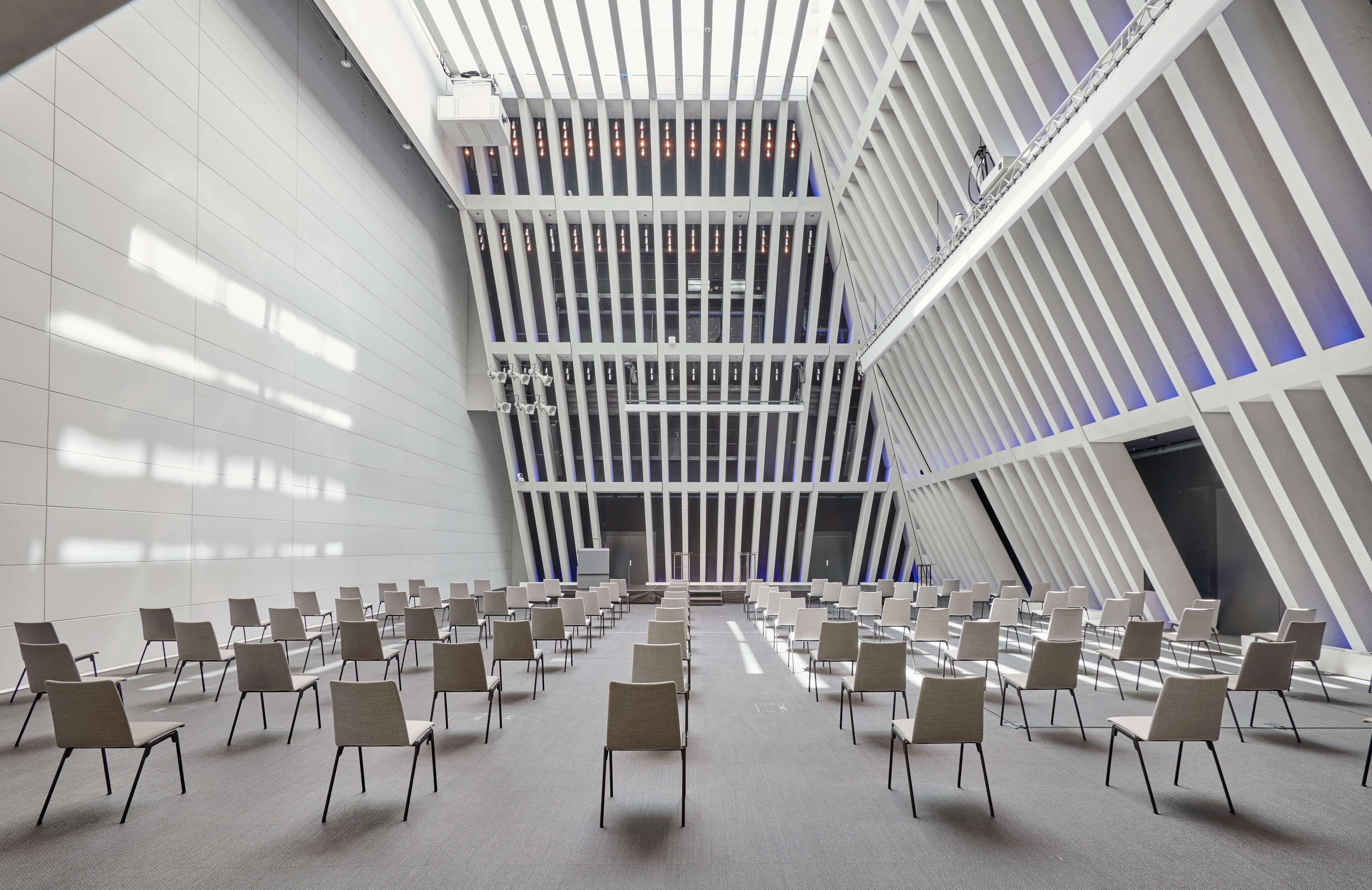 Una sala de conferencias moderna con filas de sillas orientadas hacia un escenario central, paredes blancas y un diseño geométrico en el techo.