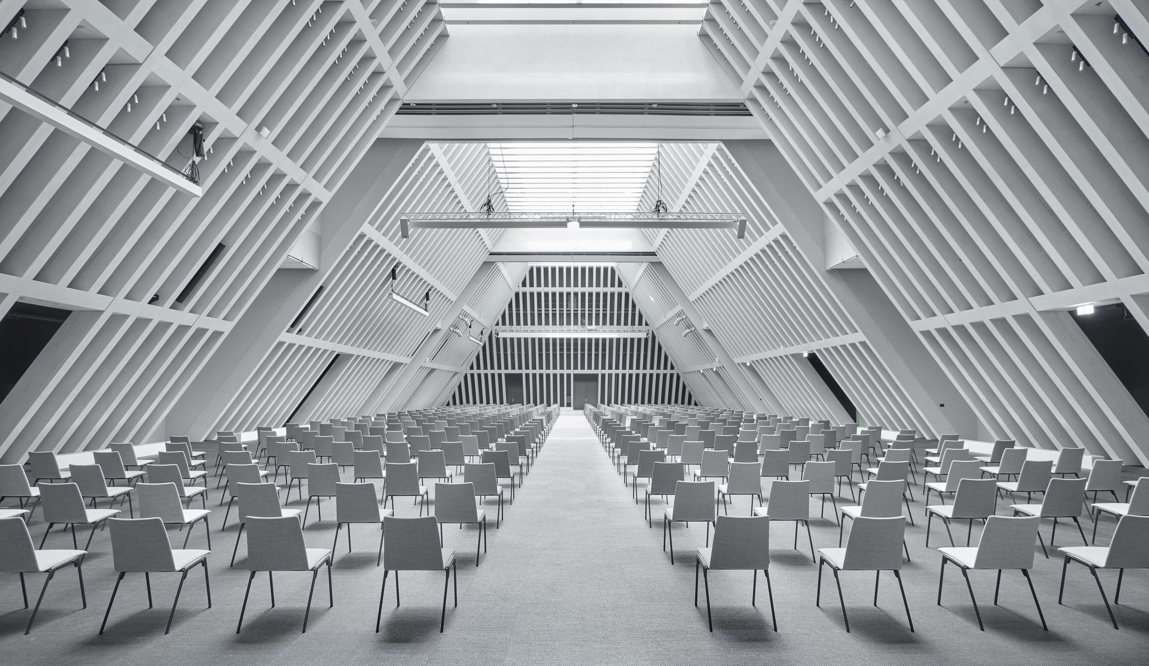 A modern conference room with rows of chairs facing a central stage, featuring geometric ceiling patterns and a symmetrical design.