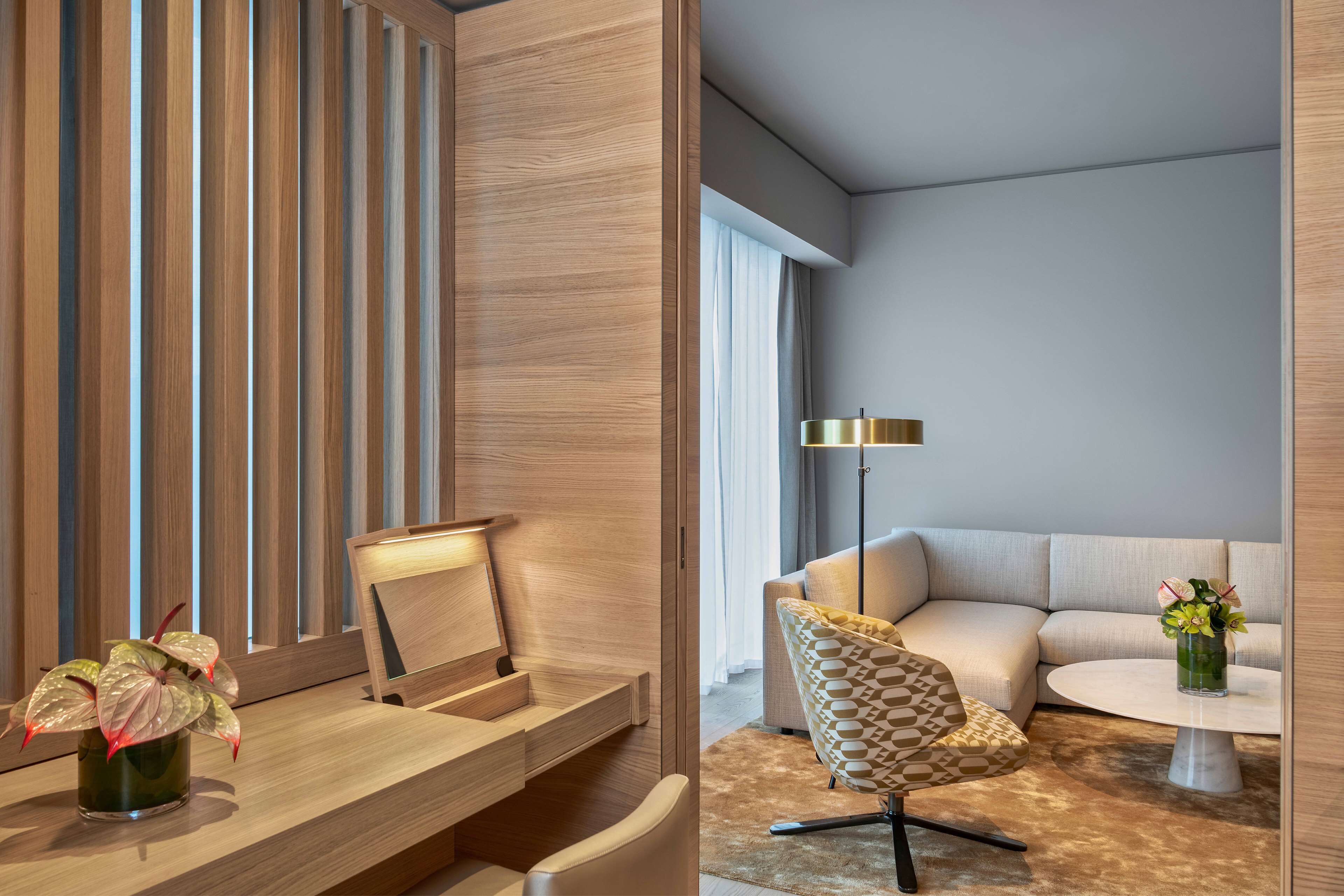 A modern hotel room with a wooden desk, a chair, a sofa, and a floor lamp.