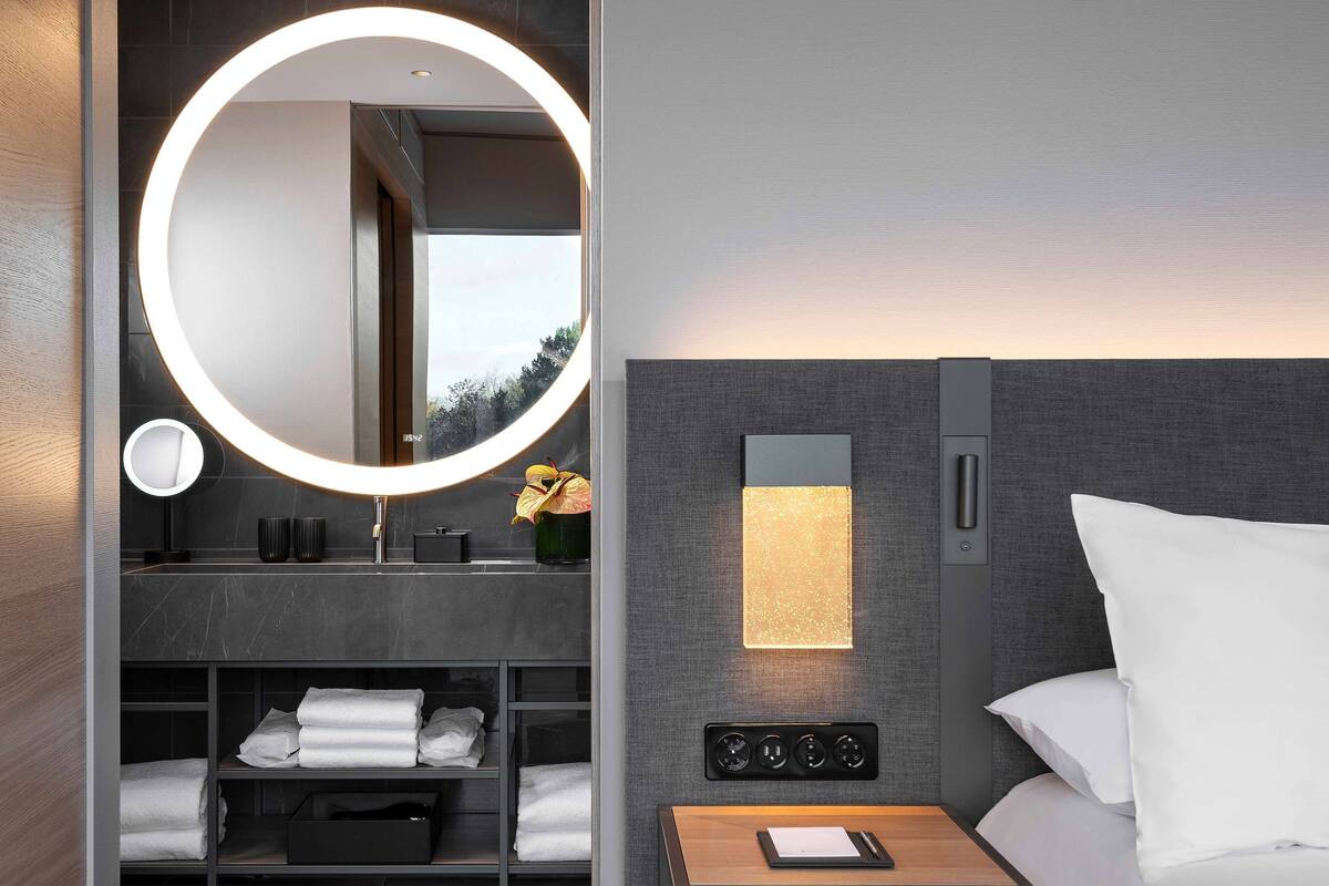 Een moderne hotelkamer met een ronde spiegel, een nachtkastje met een lamp, een wandlamp en netjes opgevouwen handdoeken.