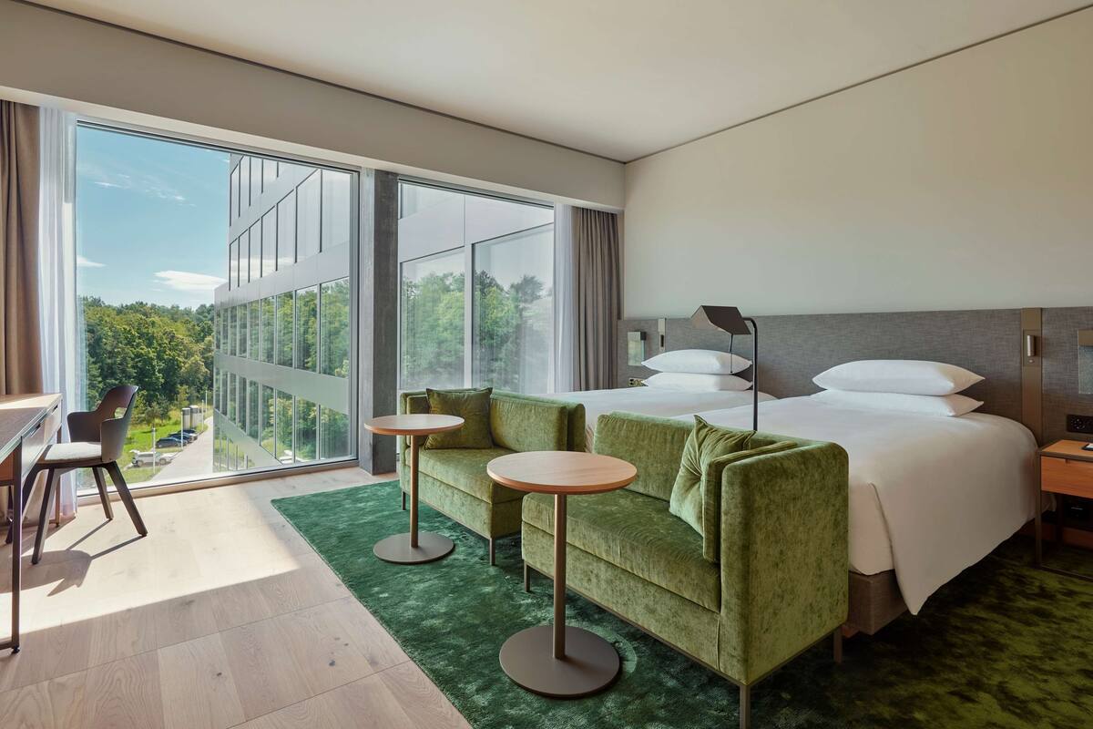Een moderne hotelkamer met een grote schuifpui naar een balkon, een groene bank, twee ronde tafels, een bed met witte lakens en een nachtkastje met een lamp.