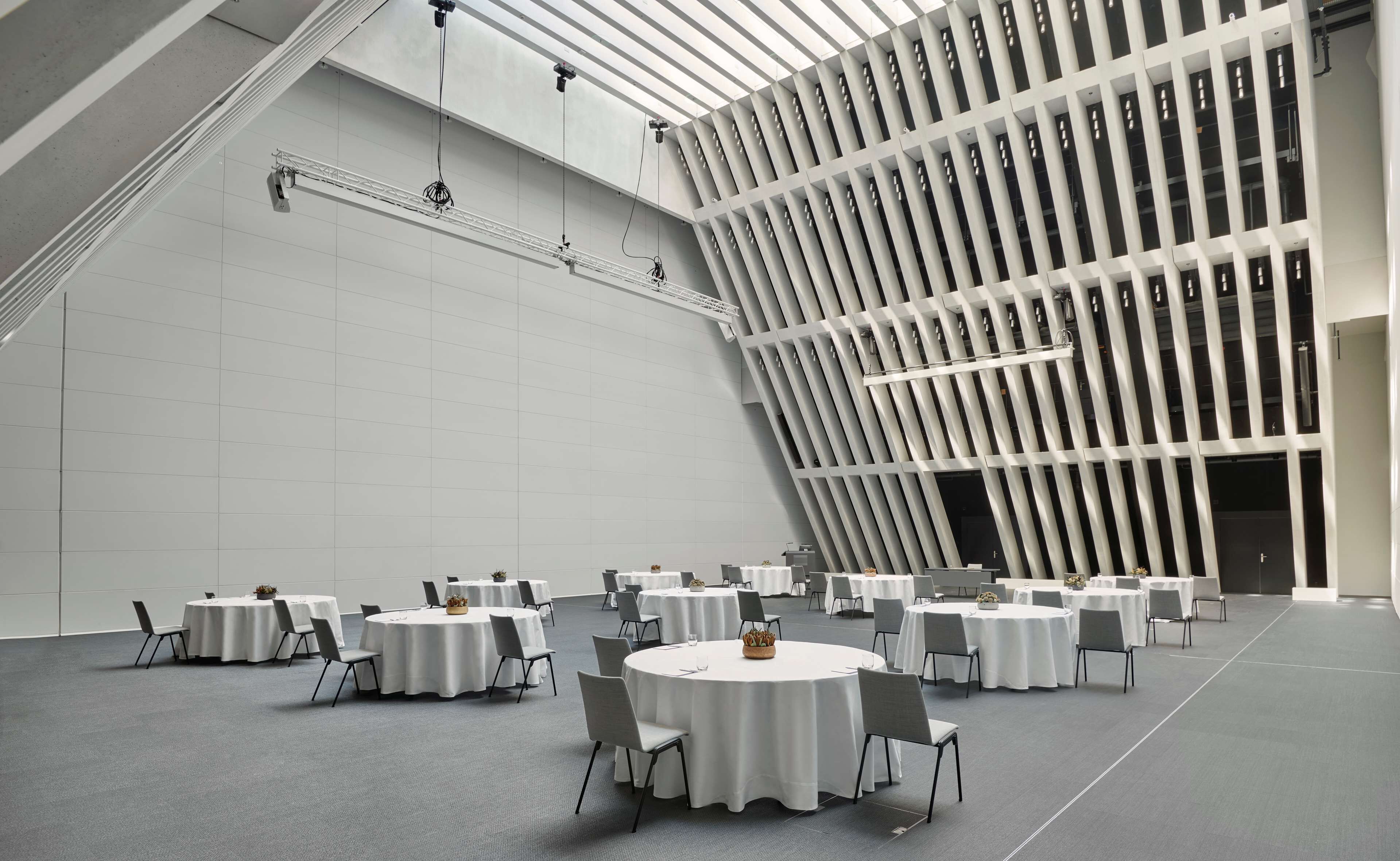 A spacious conference room with multiple round tables, grey chairs, and a modern ceiling design.