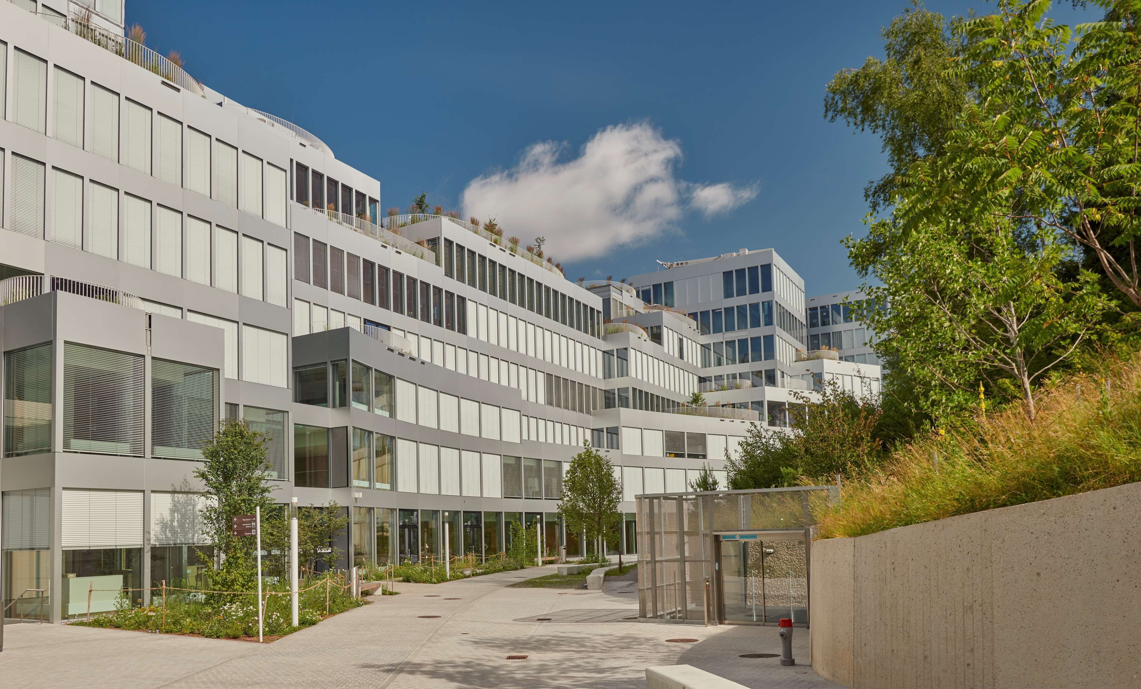 A modern multi-story building with large windows and balconies, surrounded by greenery and a clear sky.