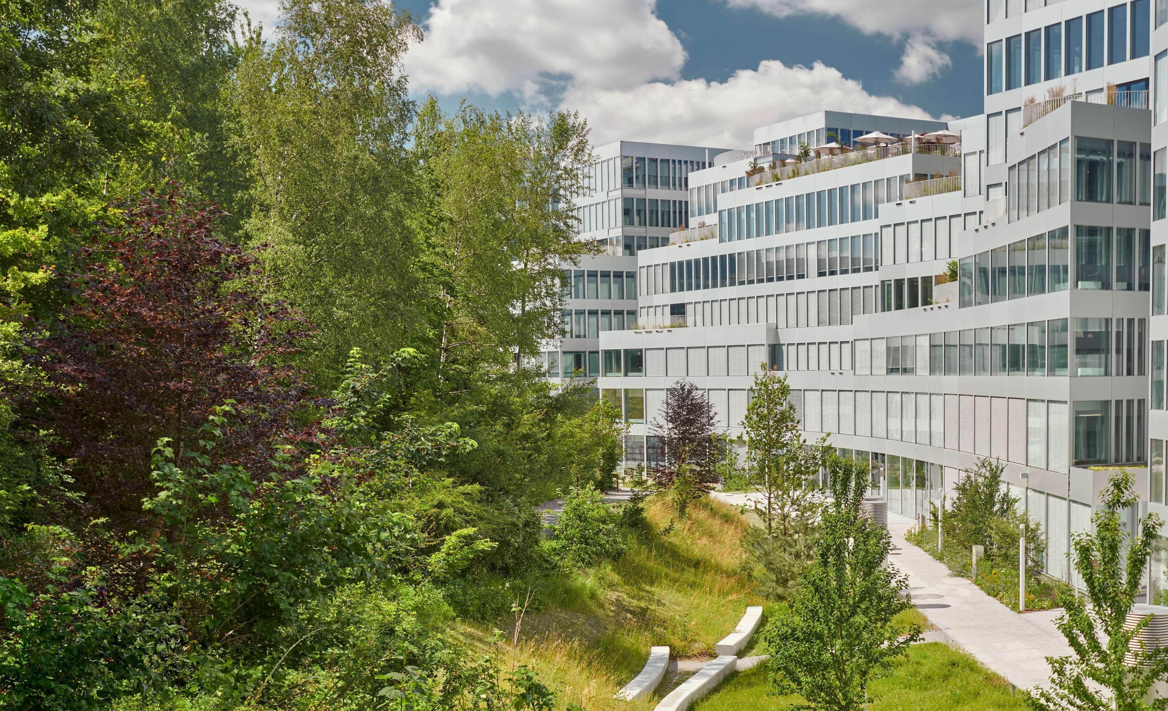 Un edificio moderno de varios pisos con amplios ventanales y balcones, rodeado de abundante vegetación y un sendero.