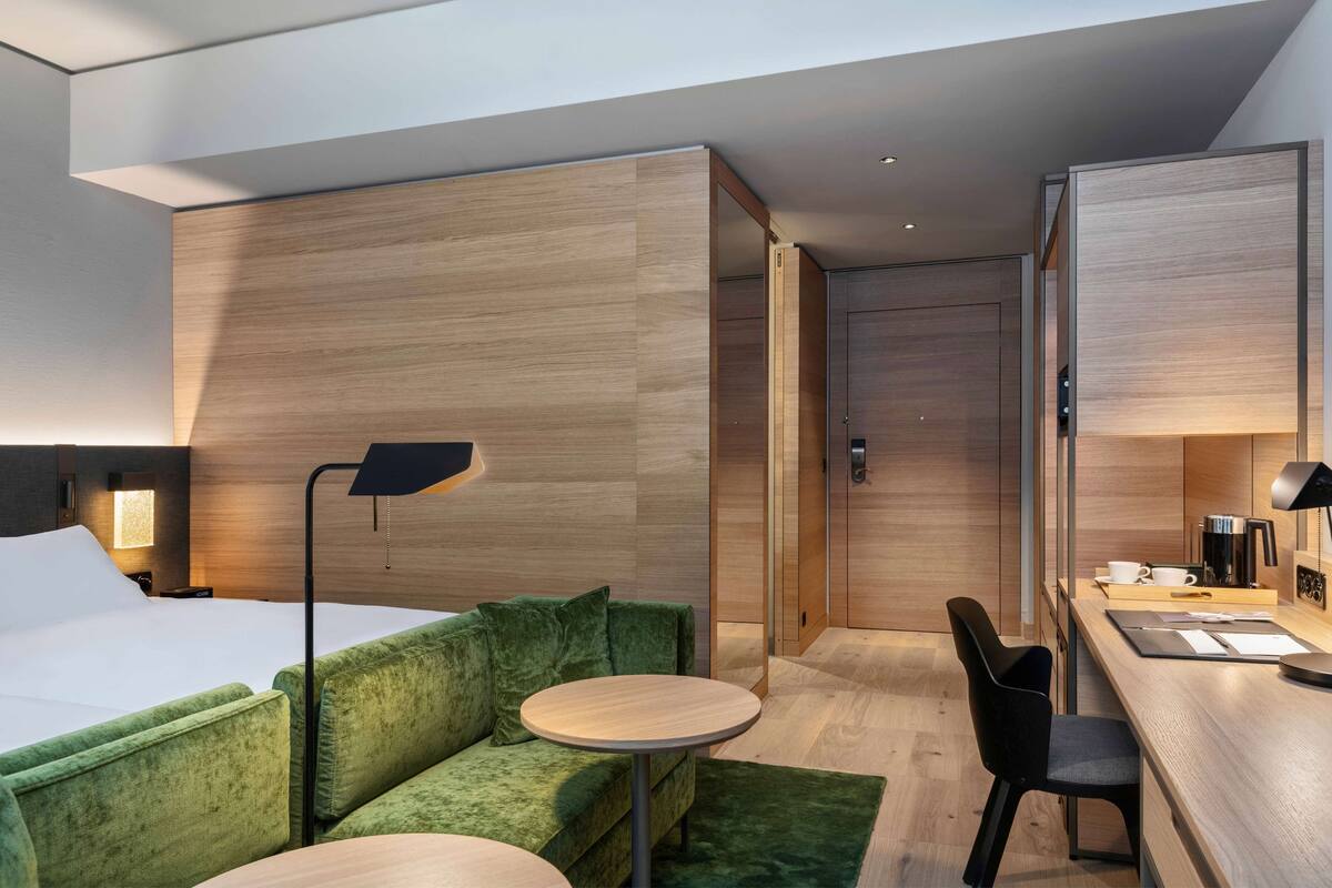 Een moderne hotelkamer met een bed, een groen gestoffeerde bank, een klein rond tafeltje, een bureau met een stoel en een spiegel.