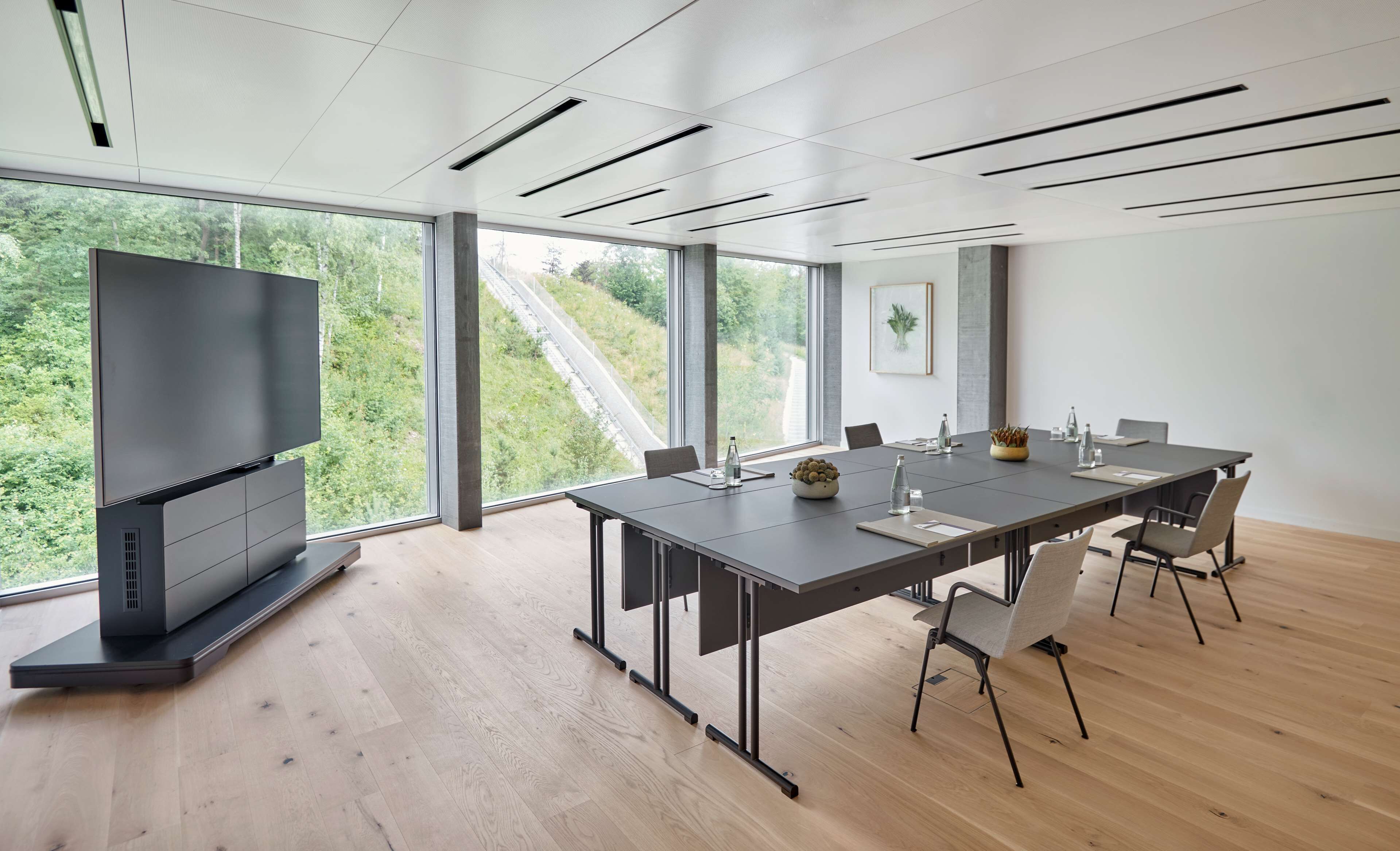 Ein moderner Konferenzraum mit langem Tisch, Stühlen, einem Flachbildfernseher und großen Fenstern mit Blick auf Grünflächen und Gleisanlagen.