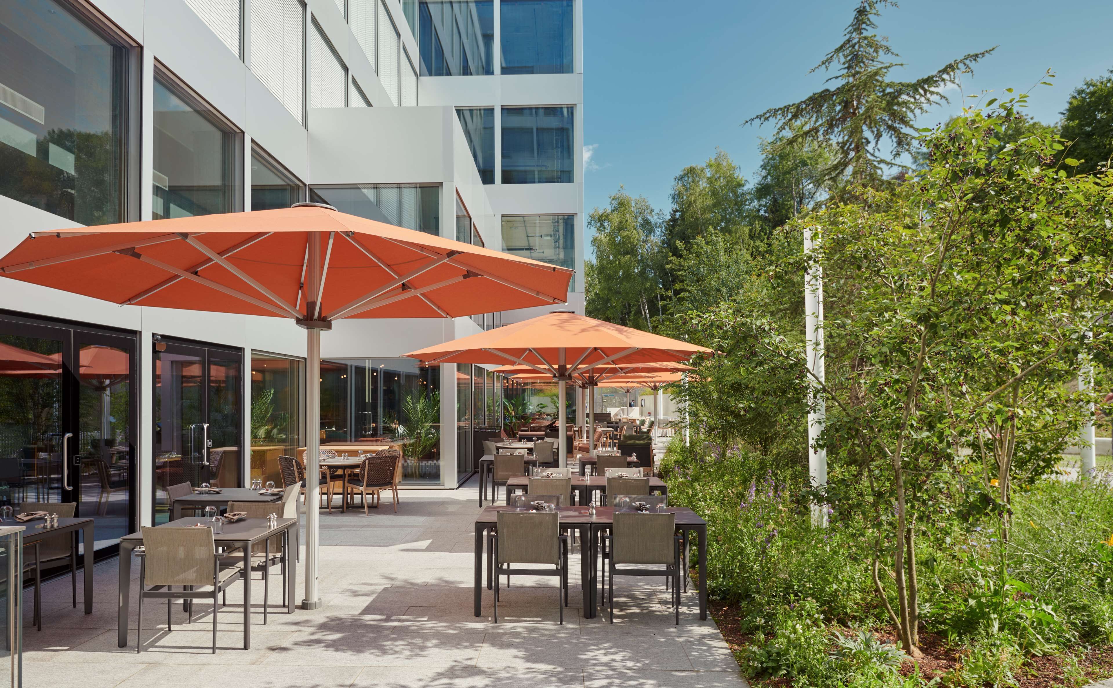An outdoor dining area with tables and chairs under orange umbrellas, surrounded by greenery and modern buildings.