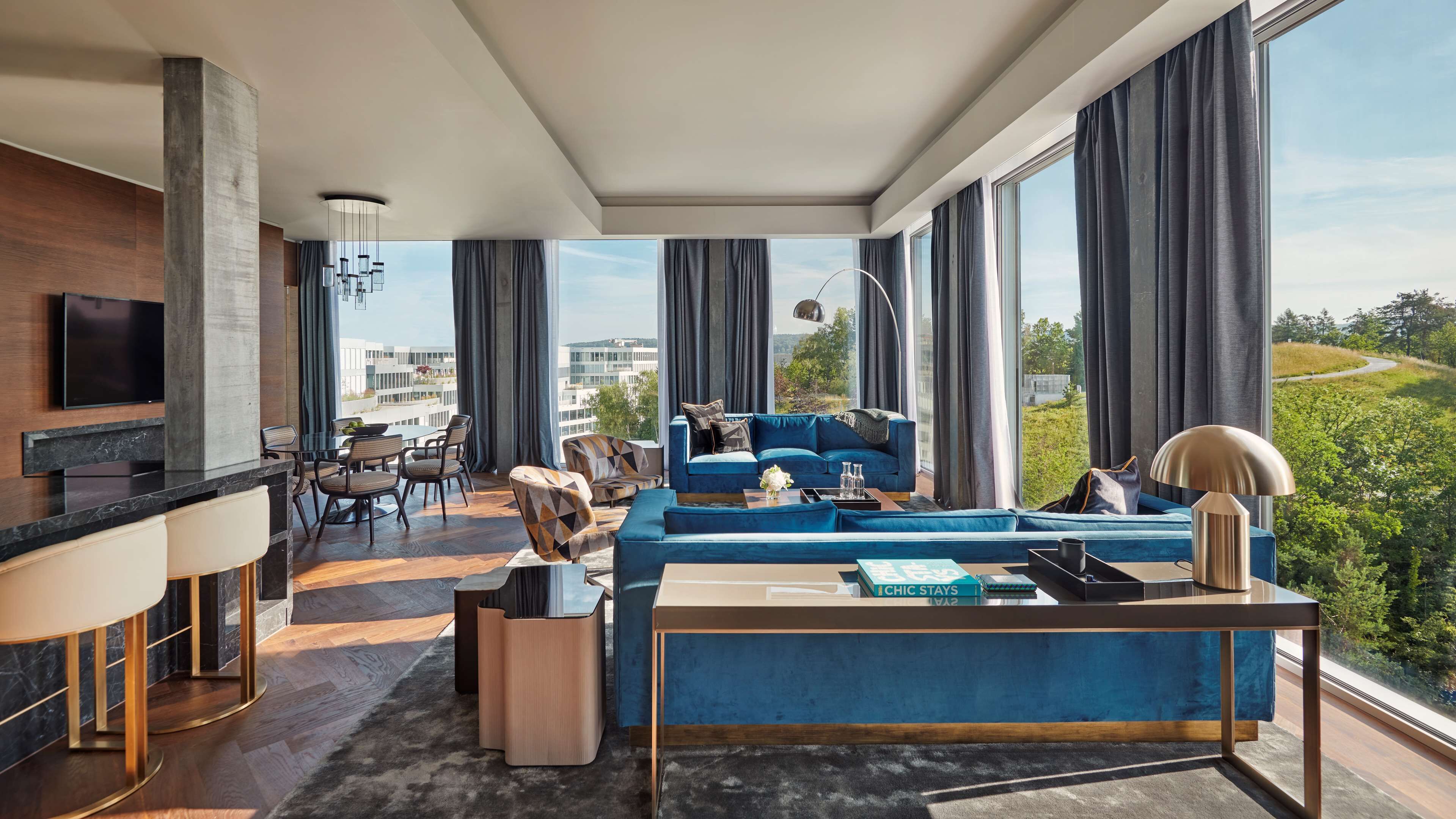Una sala moderna con un sofá azul, una mesa de centro y un televisor en la pared. Hay un comedor con una mesa y sillas, y ventanas grandes con cortinas.