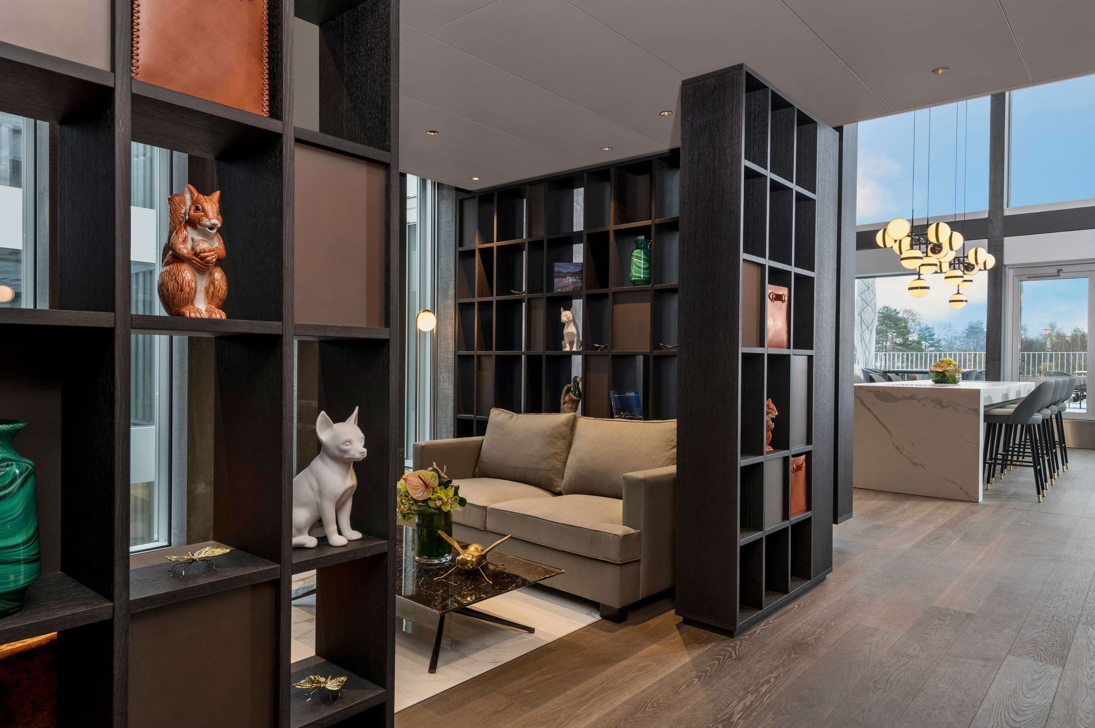 A modern living room with a sofa, coffee table, and decorative items. There is a bookshelf with various objects and a large window providing natural light.