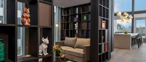 A modern living room with a sofa, coffee table, and decorative items. There is a bookshelf with various objects and a large window providing natural light.