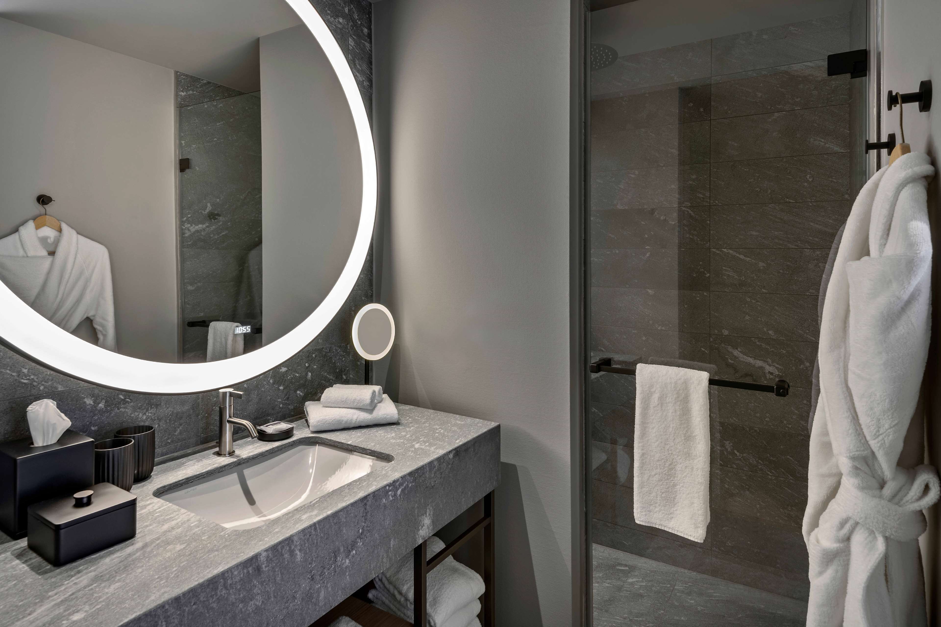 A modern bathroom with a large mirror, a round light, a sink, and a glass-enclosed shower.