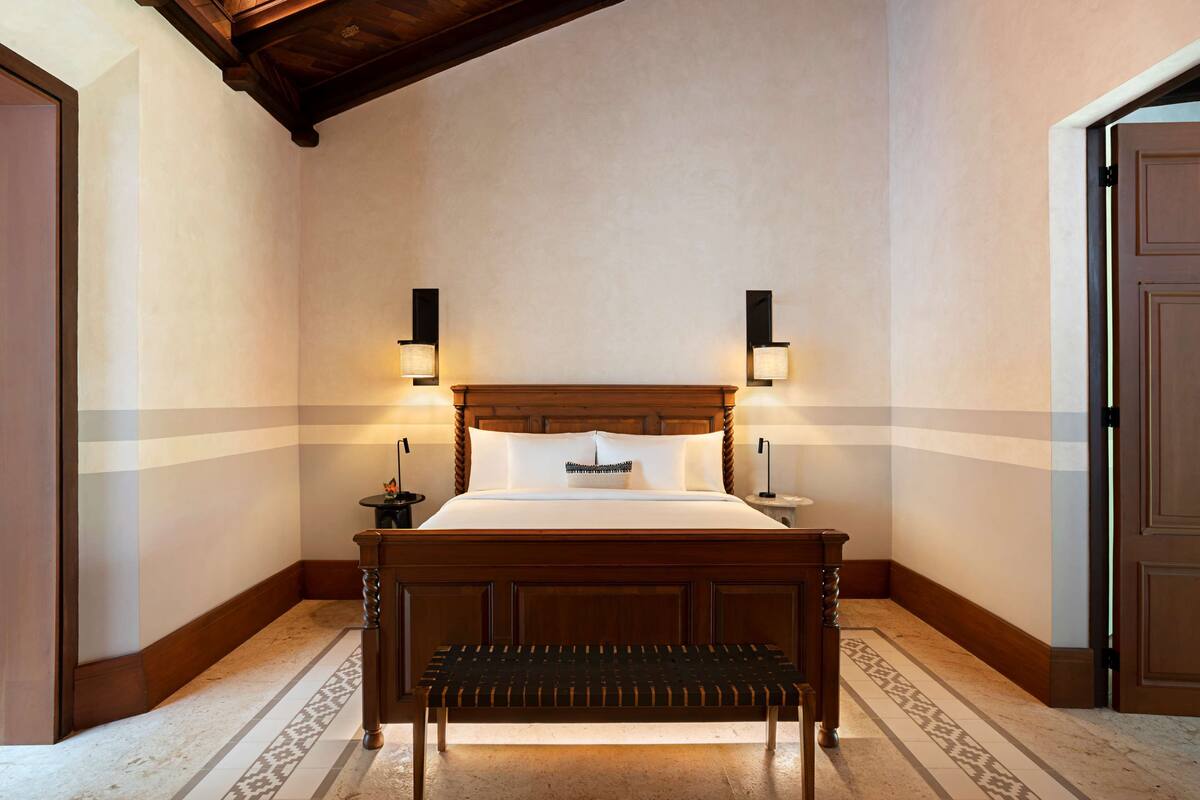 A hotel room with a wooden bed, two bedside lamps, a bench, and a patterned floor.