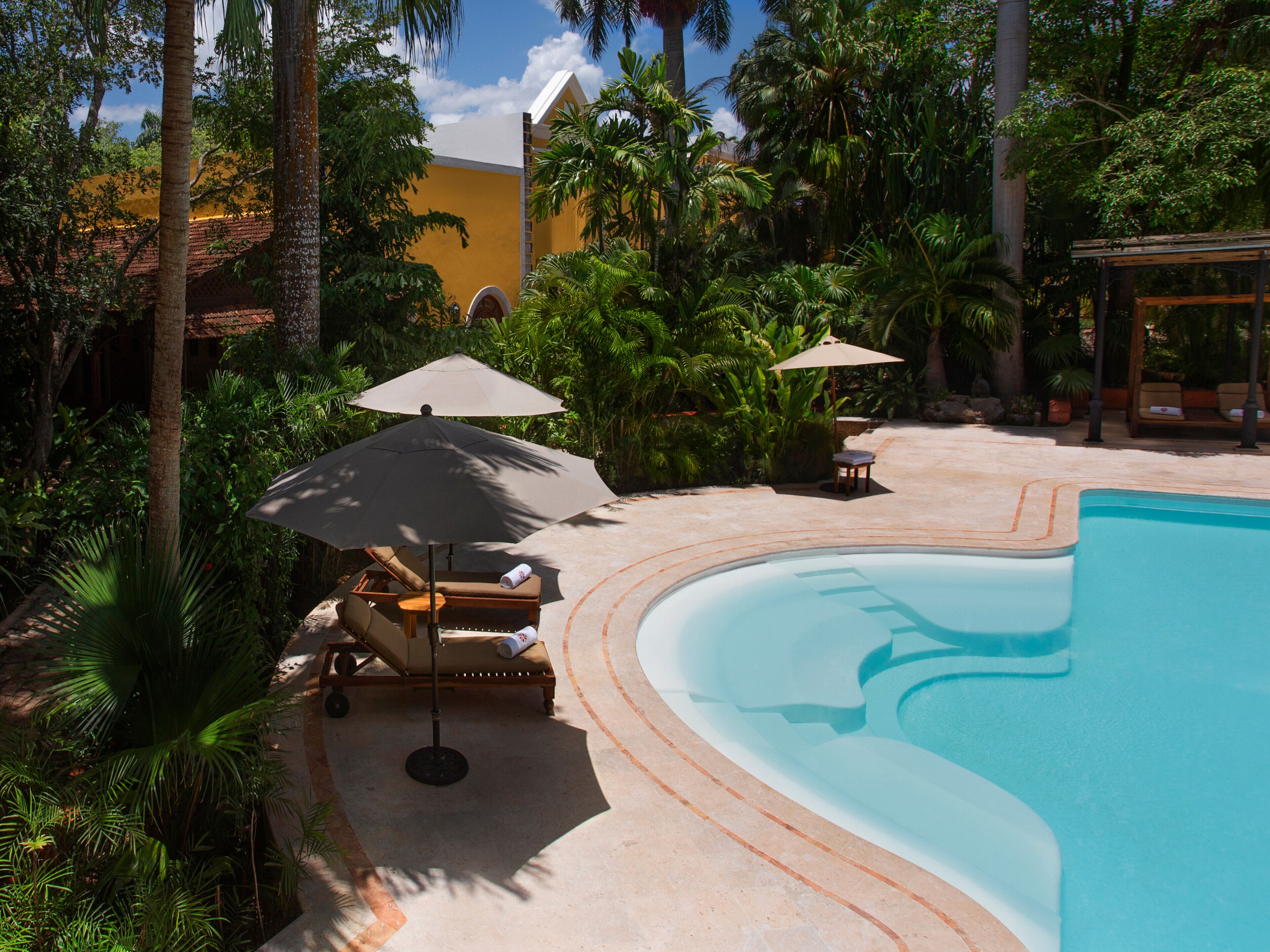 A pool area with lounge chairs and umbrellas, surrounded by palm trees and a building with a yellow facade.