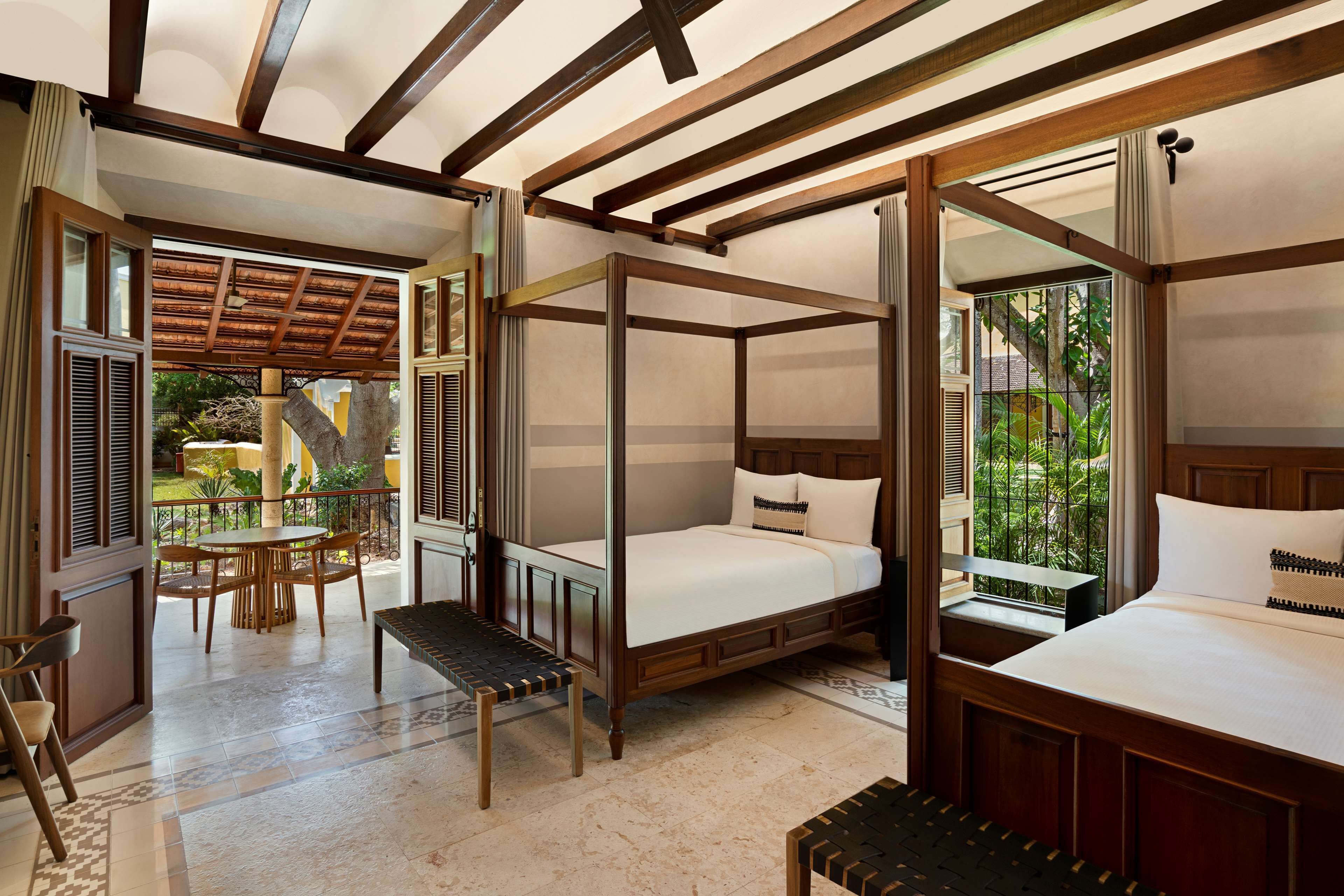 A spacious bedroom with a large bed, a smaller bed, and a bench. There is a balcony with a view of greenery.