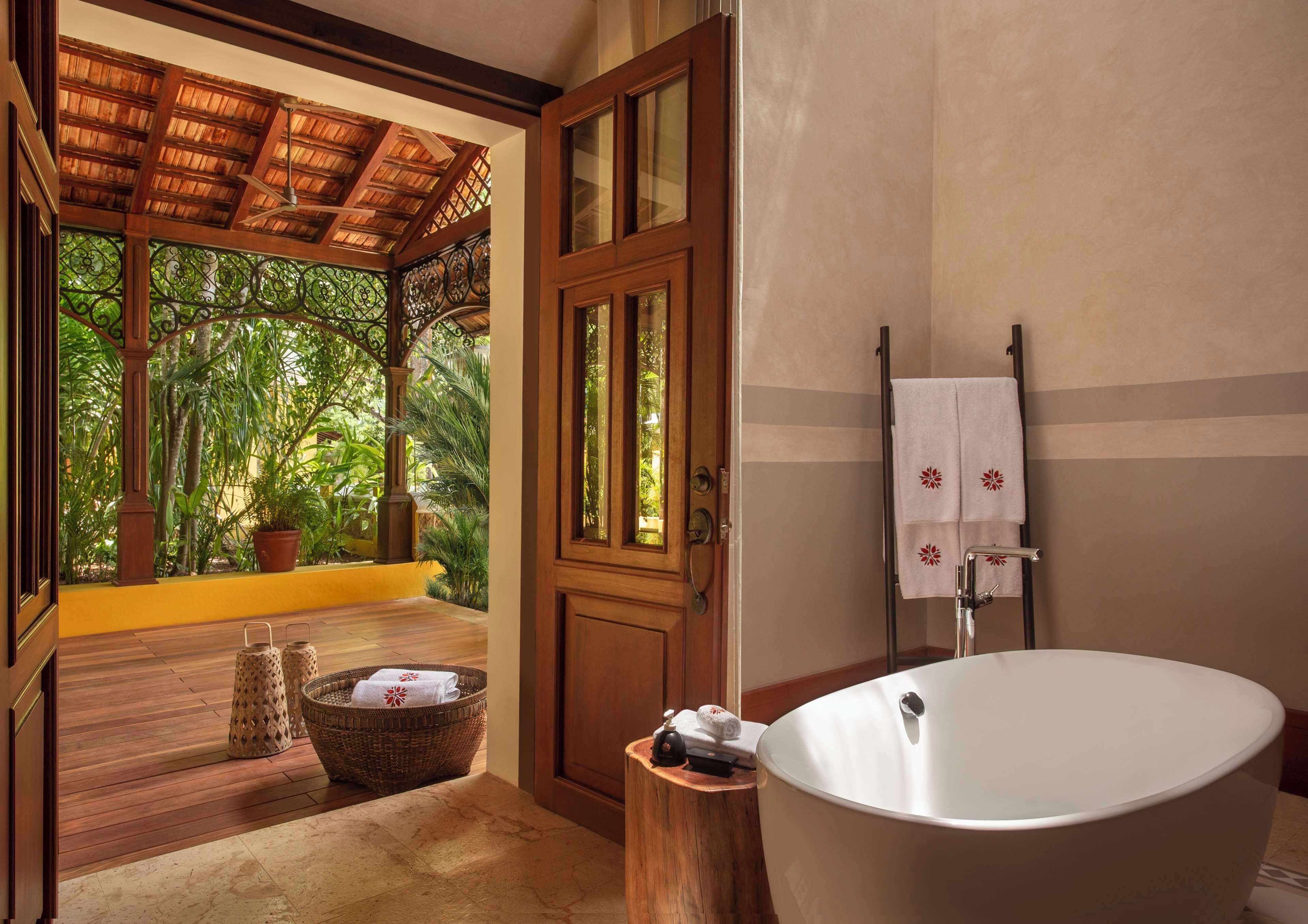 A bathroom with a freestanding bathtub, a wooden door, and a view of lush greenery through a window.