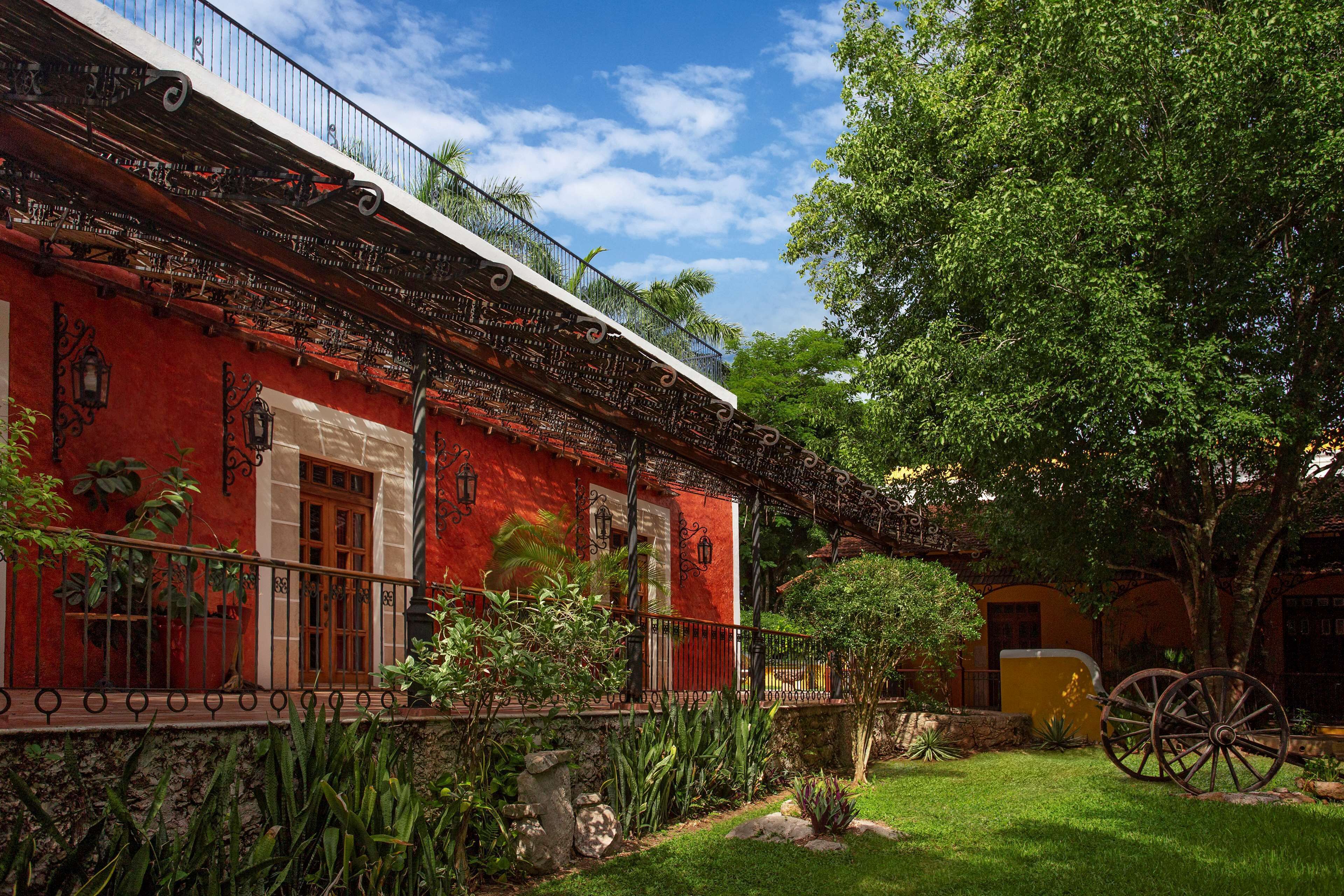 A red building with a balcony, surrounded by greenery and a large tree.