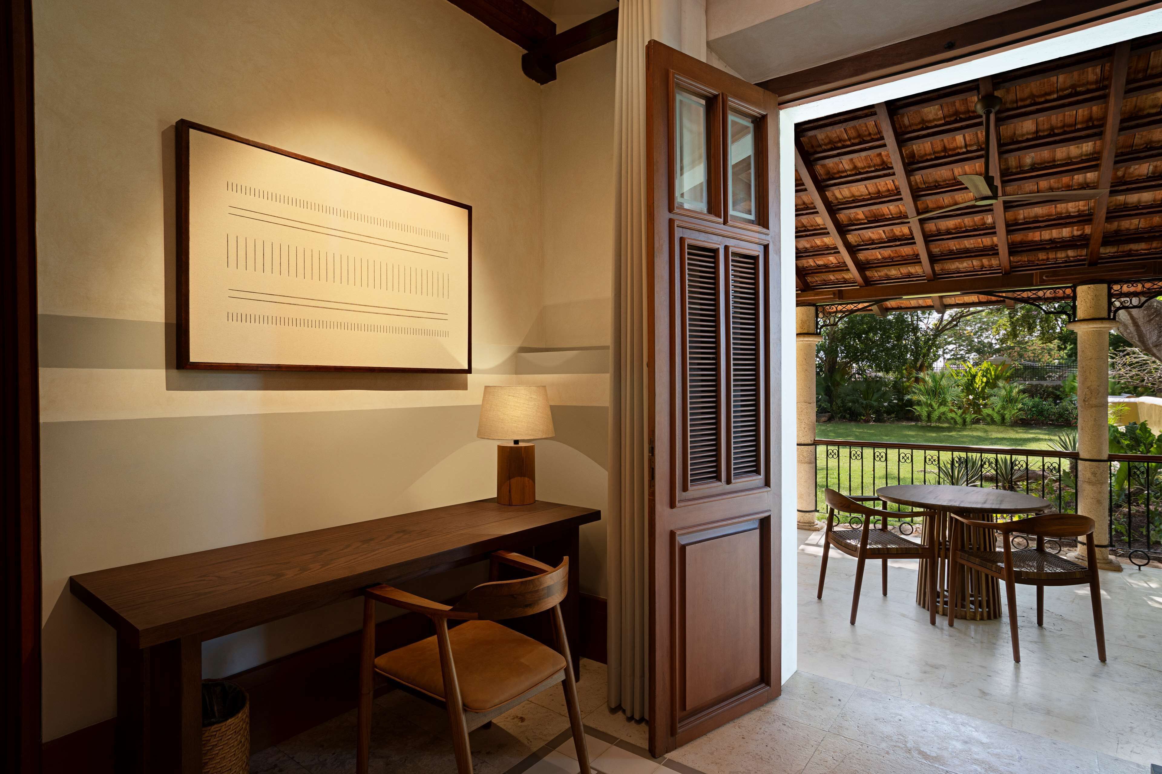 A room with a wooden desk, a chair, a framed picture, and a view of a garden through a window.