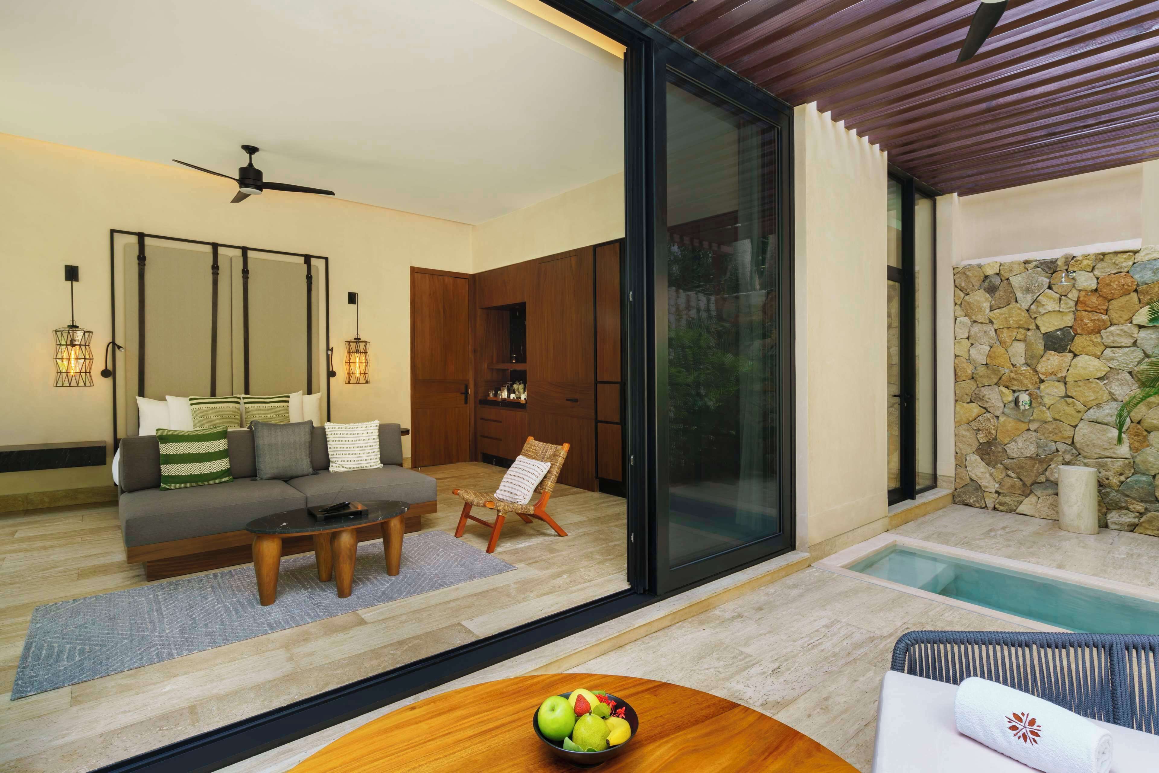 Sala de estar moderna com sofá, mesa de centro e mesa de jantar com frutas. Há uma porta de vidro que dá para uma área de piscina com parede de pedra.