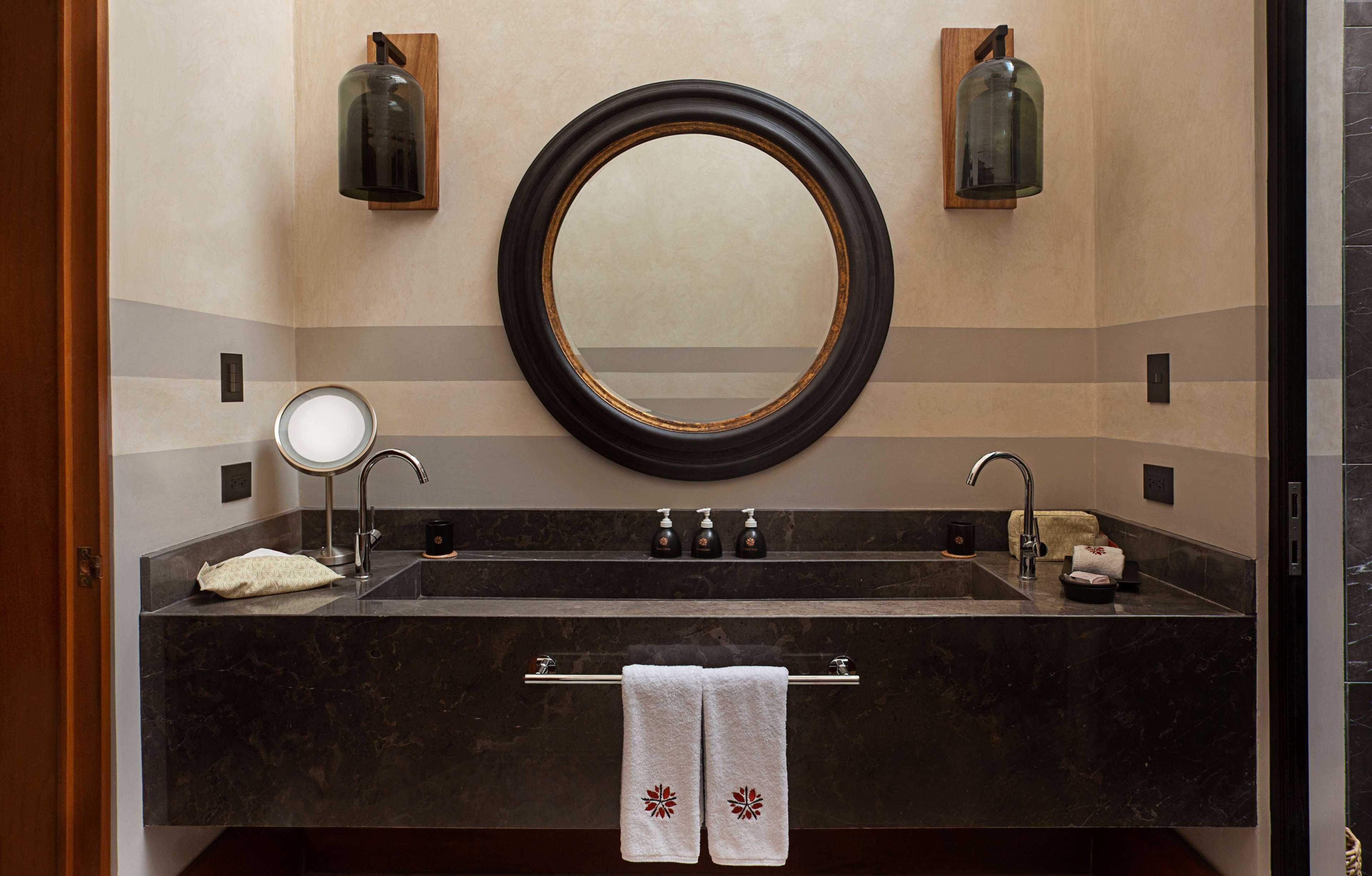 A bathroom with a dark marble sink, two faucets, a round mirror, and two wall-mounted light fixtures.