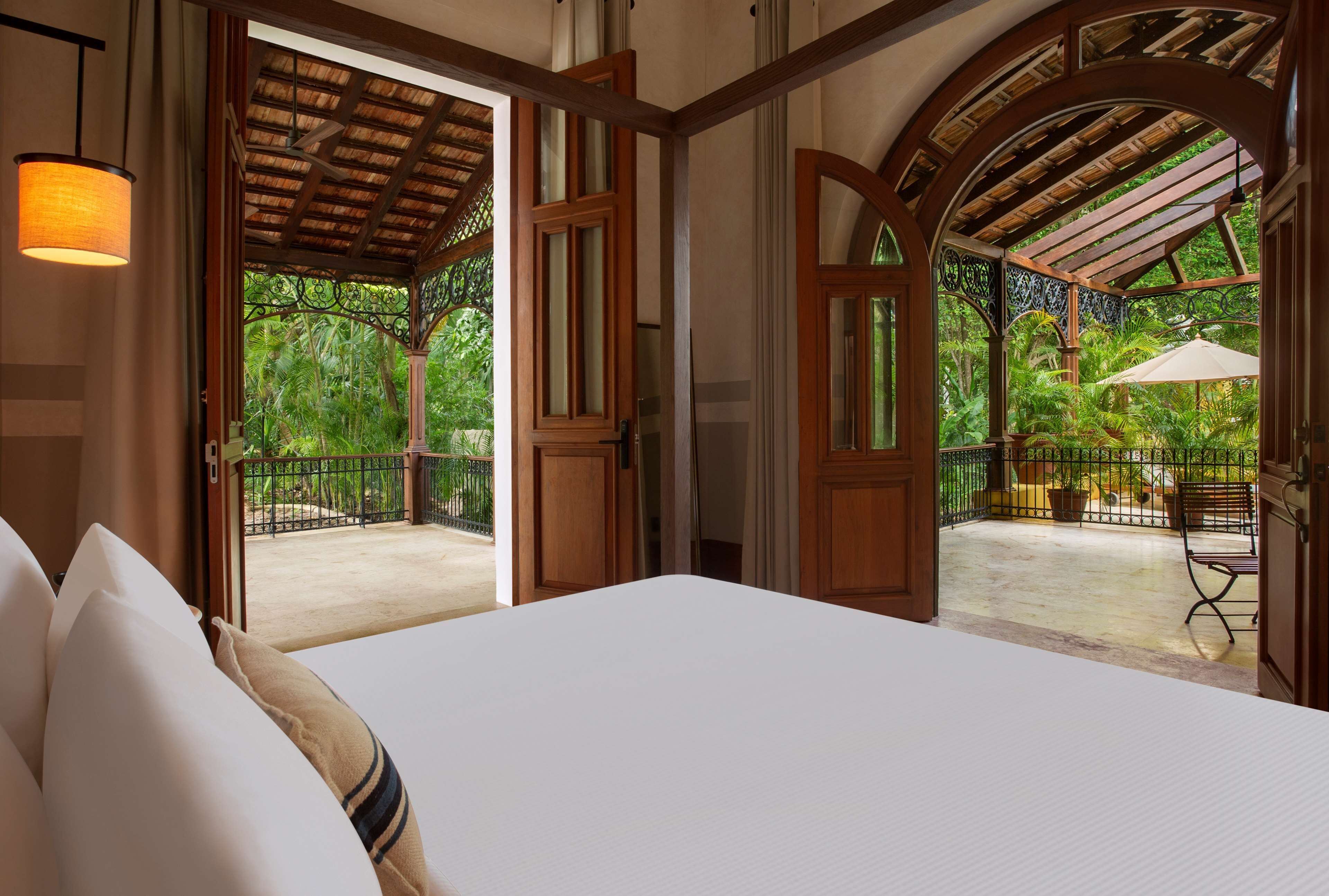 A hotel room with a large bed, wooden doors leading to a balcony with a view of lush greenery.