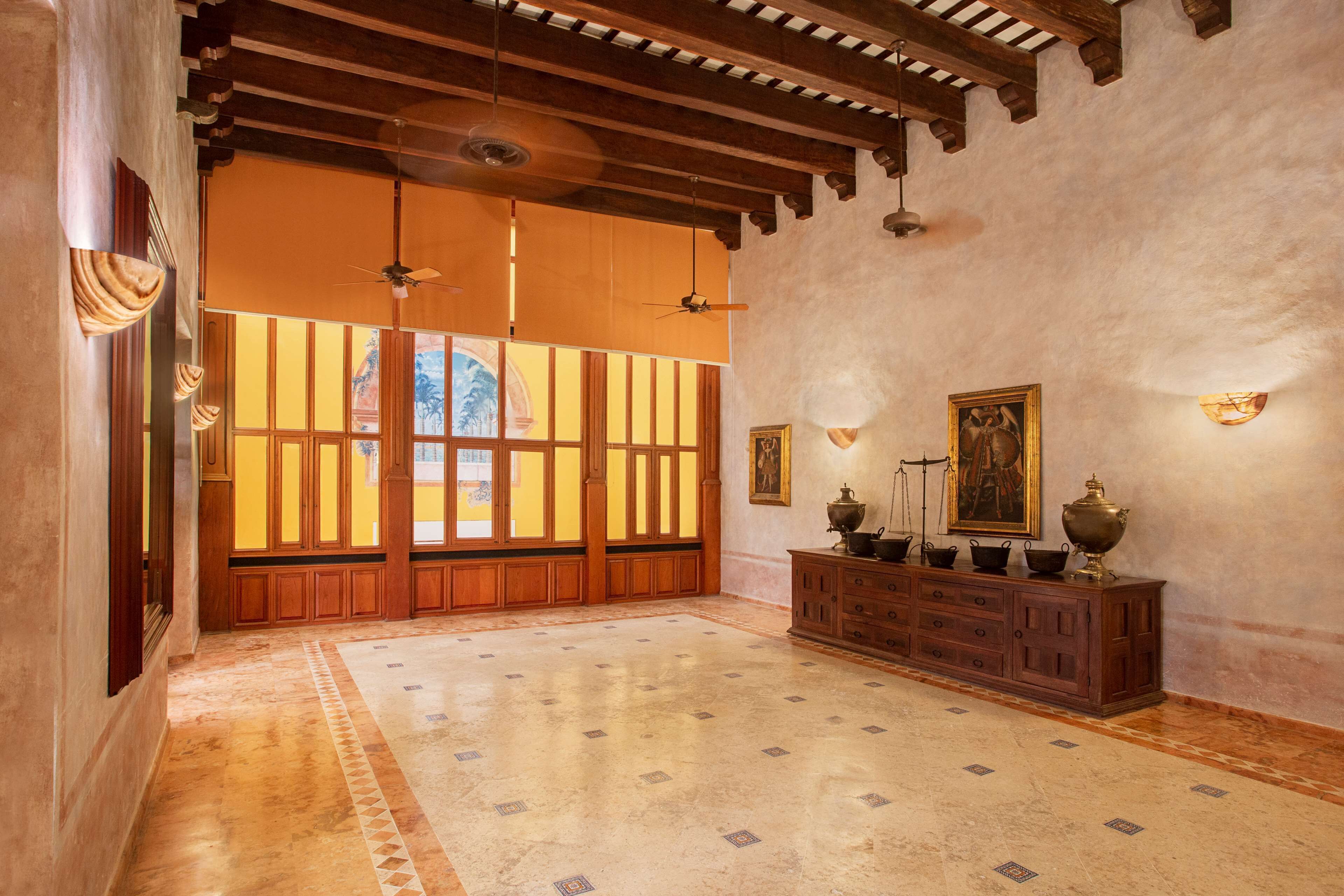 A spacious room with a high ceiling, wooden beams, a tiled floor, a large window with vertical blinds, and a wooden cabinet with decorative items.