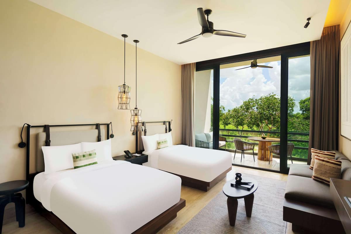 A hotel room with two beds, a ceiling fan, a balcony with a table and chairs, and a view of greenery.