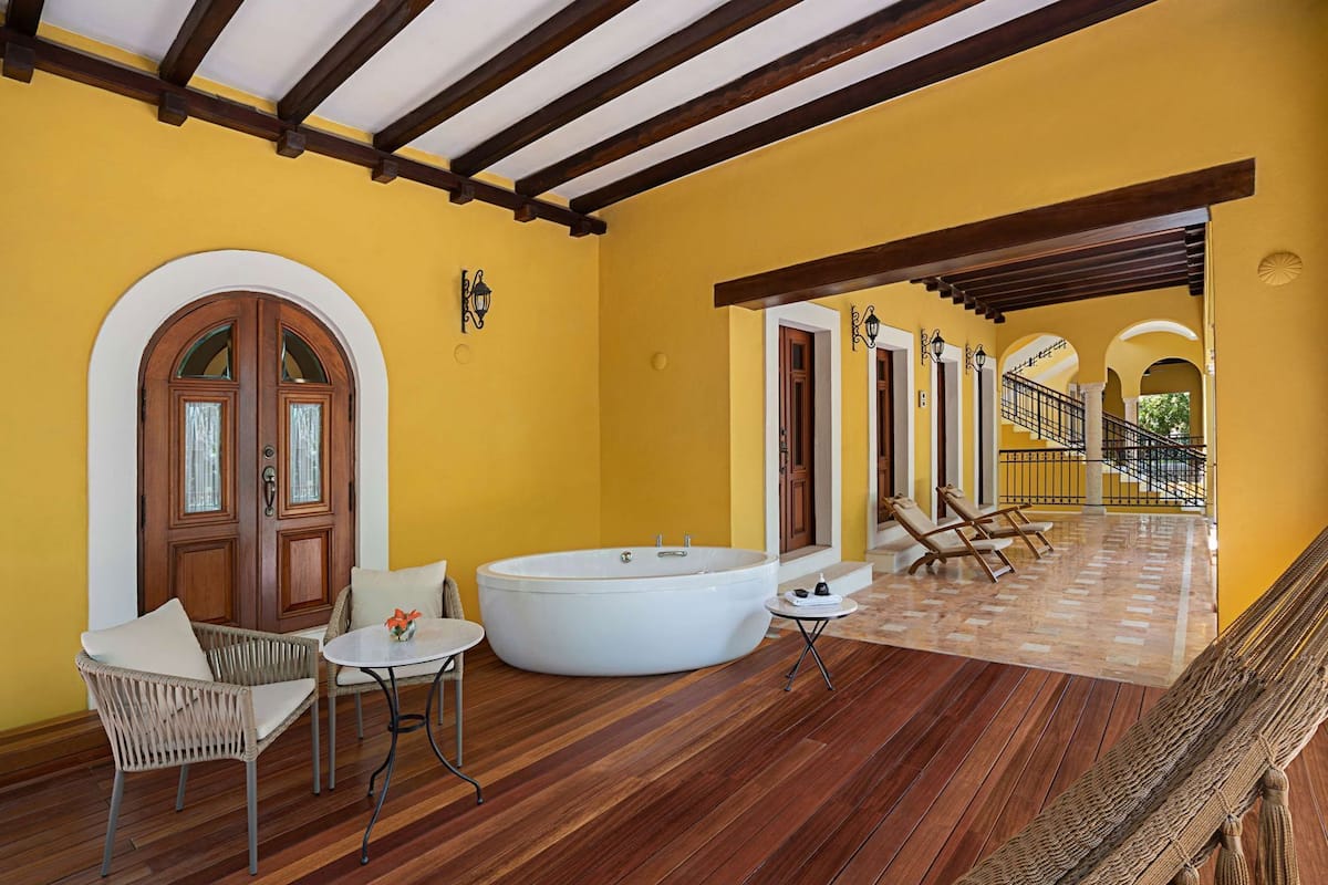 A yellow-walled room with a large bathtub, wicker chairs, and a wooden floor.