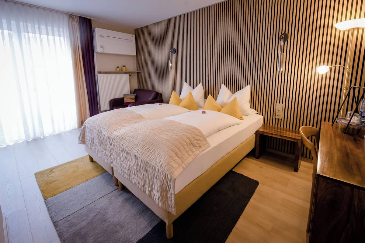A modern hotel room with a large bed, a wooden headboard, a desk with a chair, and a window with curtains.
