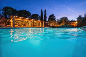 A swimming pool with a covered area and lounge chairs, surrounded by trees and a building with a lighted facade. - Farmhouse Apartment, Tournesol 6+1, Volterra Area, San Gimignano, Pisa (Montelopio di Peccioli)