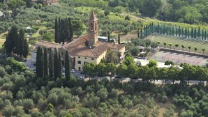 Oversigtsbillede af en kirke med et klokketårn, omgivet af oliventræer og en tennisbane.