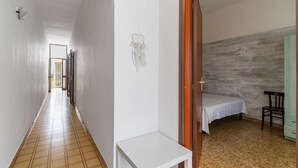 Un couloir doté d’un sol carrelé, d’un mur blanc et d’une porte donnant sur une pièce où se trouvent un lit et une chaise.