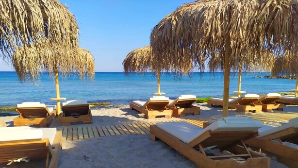 A beach with wooden lounge chairs and thatched umbrellas, arranged on a sandy area with a view of the sea. - Silver Beach Hotel (Kos)