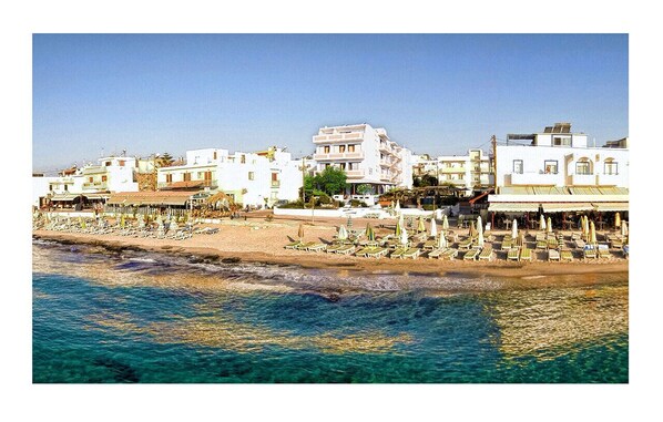 Un pueblo costero con una playa bordeada por sillas de playa y sombrillas, edificios al fondo y agua azul cristalina.