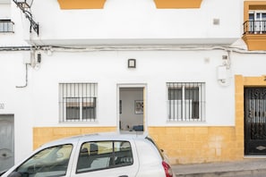 A white building with a yellow lower section, a car parked in front, and a black gate to the right.