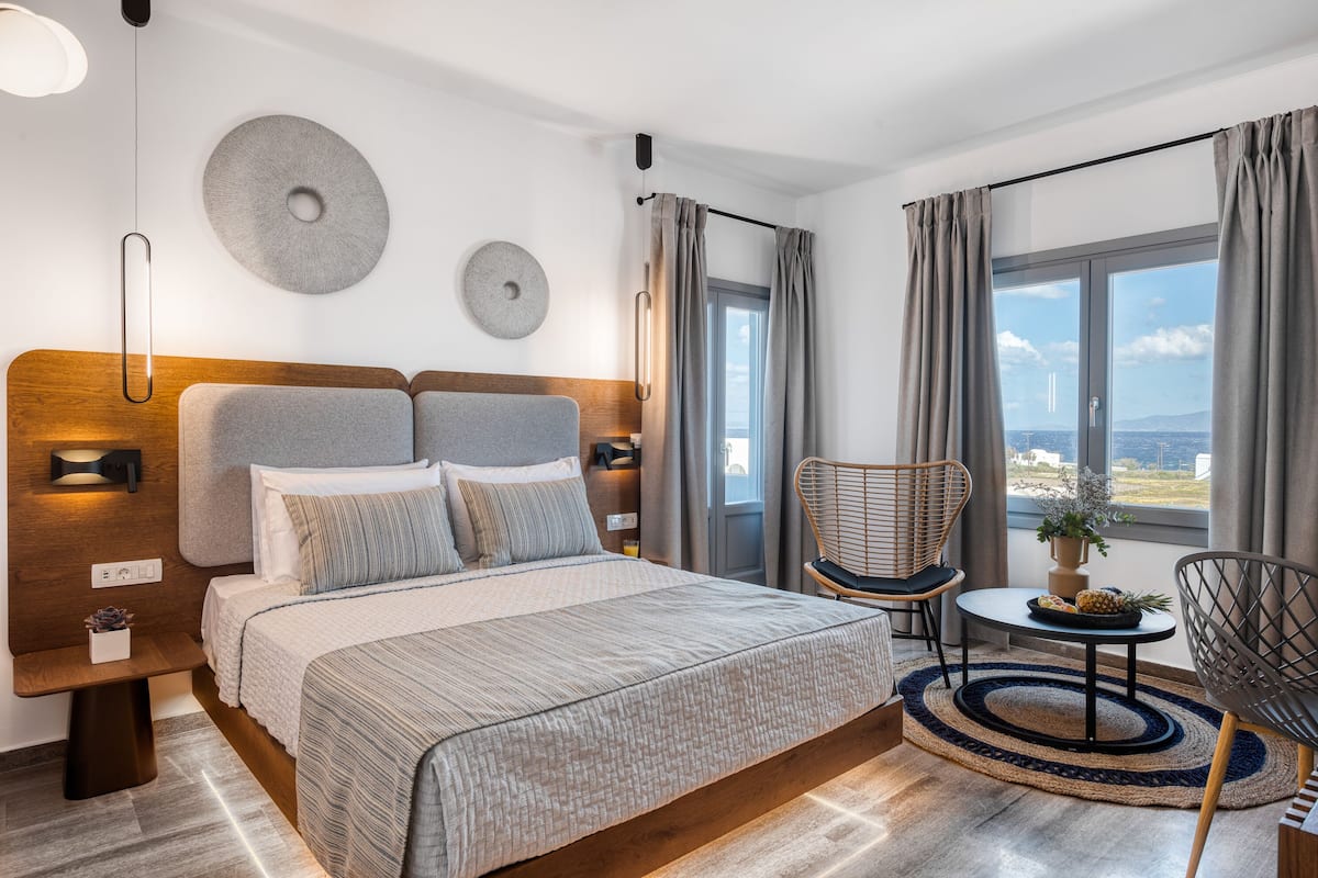 Quarto moderno com uma cama grande, cabeceira de madeira, mesa lateral, uma cadeira, pequena mesa com uma planta e vista da cidade por grandes janelas.