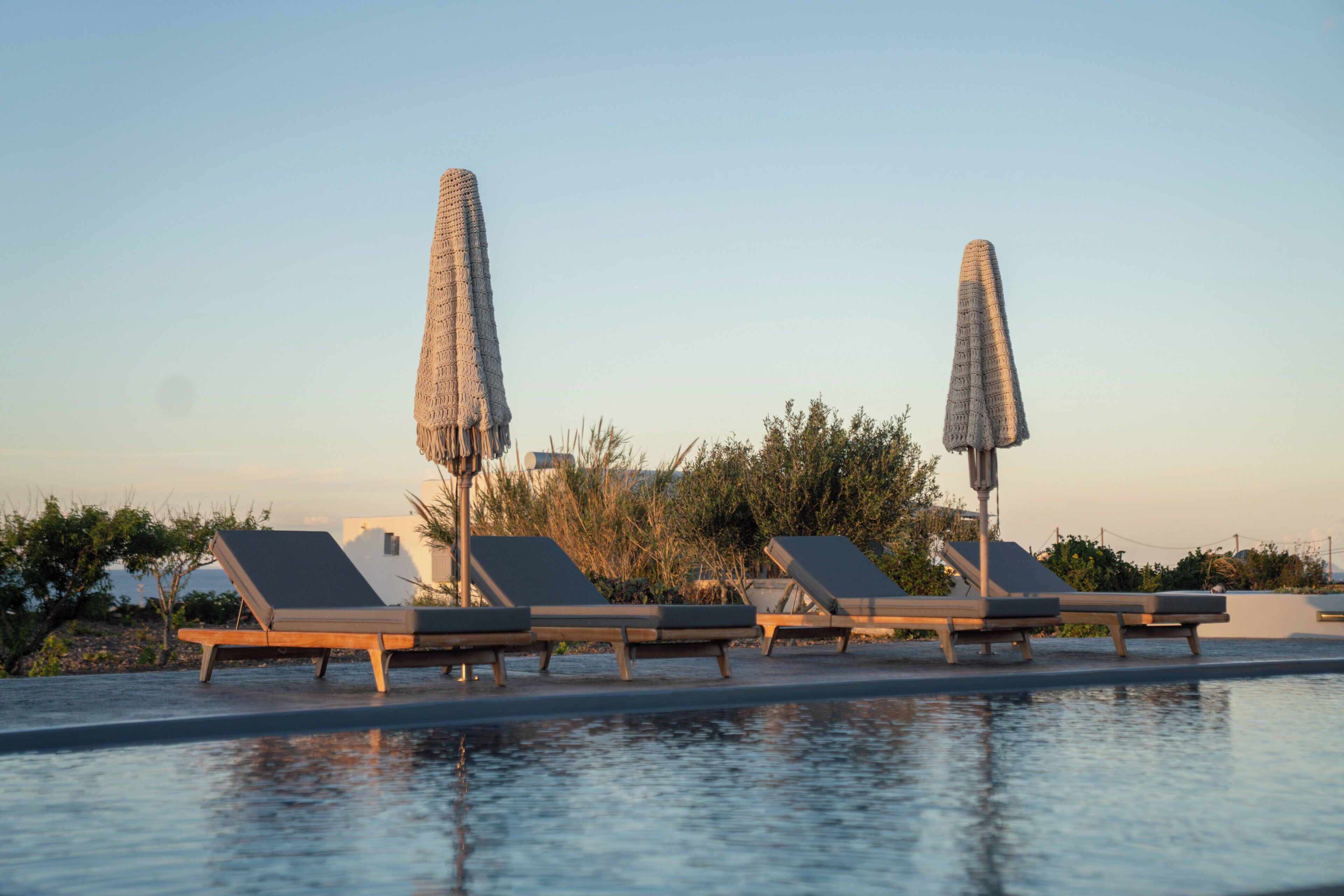 Zona junto a la piscina con sillones de descanso y sombrillas, cielo despejado y vegetación al fondo.