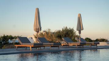 Zona junto a la piscina con sillones de descanso y sombrillas, cielo despejado y vegetación al fondo.