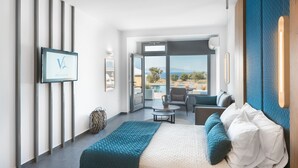 A modern hotel room with a bed, a seating area with a table, and a large window offering a view of the outdoors. - Volcanic Arc Suites (Santorini)