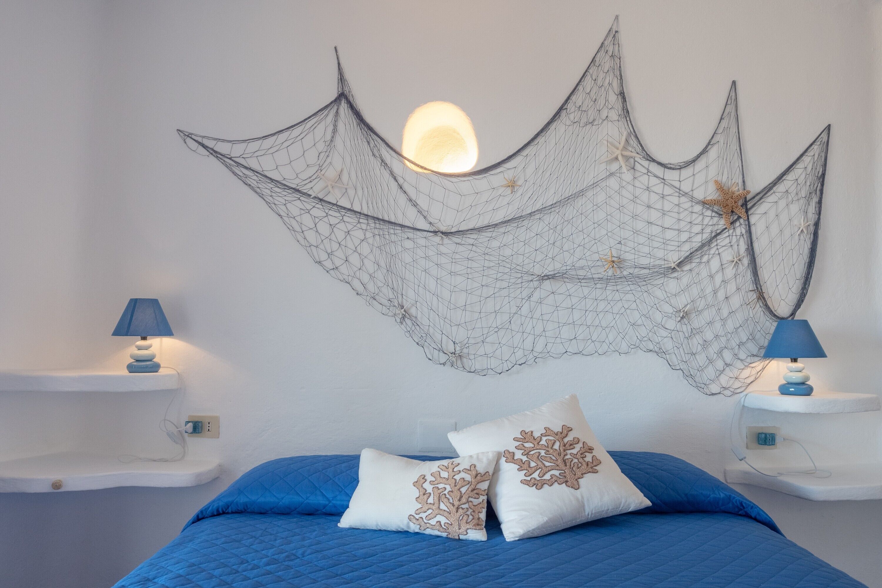 Un lit avec une literie bleue, deux tables de chevet blanches avec des lampes, et une décoration murale en forme de filet.