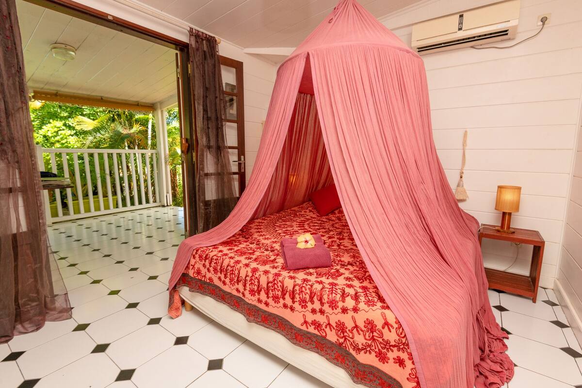 A room with a bed under a pink canopy, a bedside table with a lamp, and a view of a balcony with a white fence and greenery.