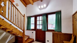 Una escalera de madera que lleva a un rellano, una puerta de madera con un ventanal y una silla en una habitación con paredes blancas y suelo de baldosas.